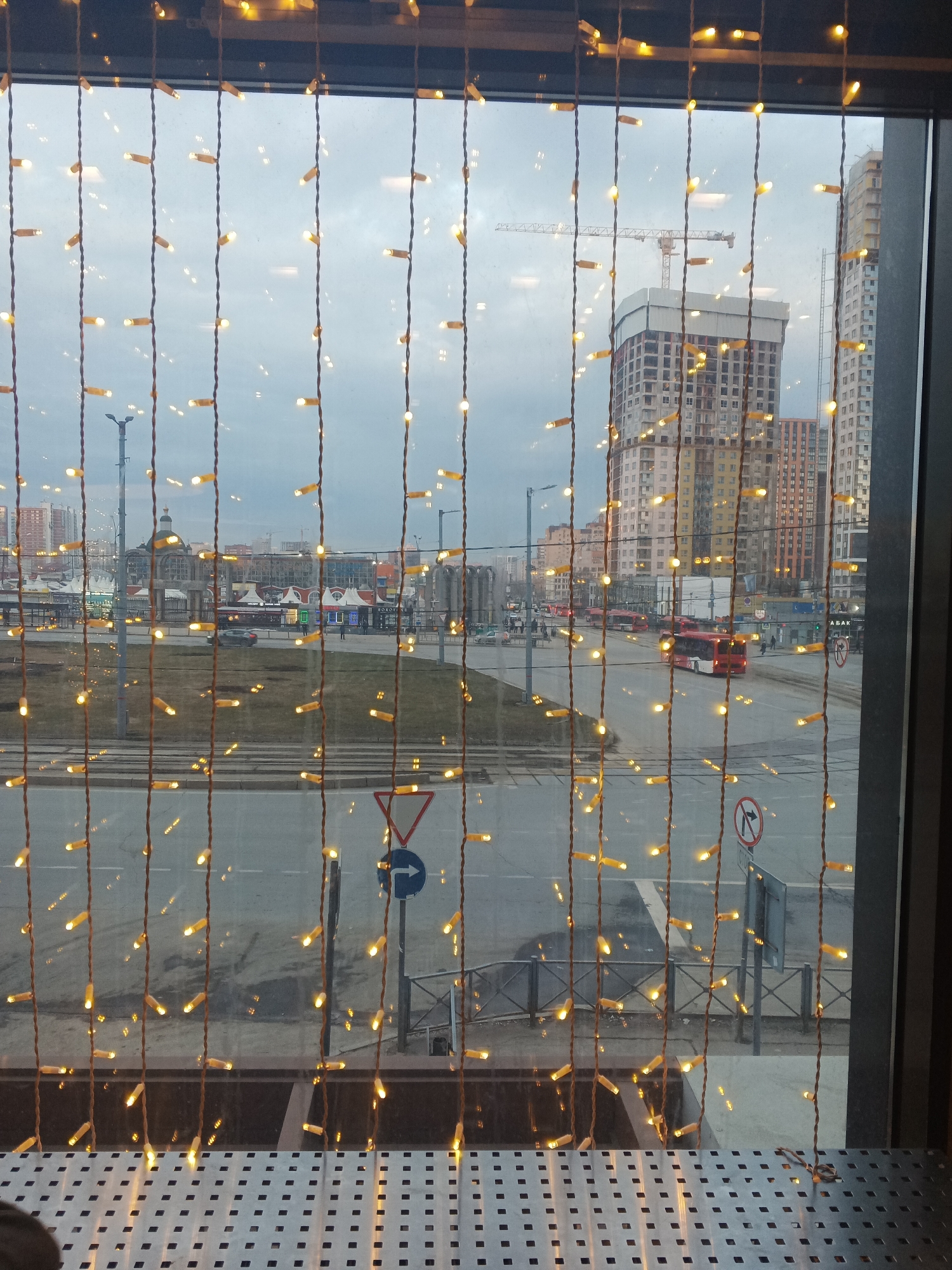 The image shows a view through a window with hanging string lights, likely inside a building, onto a city street scene. The location is Perm, Russia, as evidenced by the architectural styles of the buildings and the public transportation (red buses). The scene includes a multi-lane road with pedestrian crossings, a roundabout, and various street signs. Several tall buildings are visible in the background, some under construction, with a crane present. The sky is overcast. In the foreground, there's a perforated metal surface, probably a tabletop or counter. There's minimal activity; a few vehicles and pedestrians are visible in the distance.