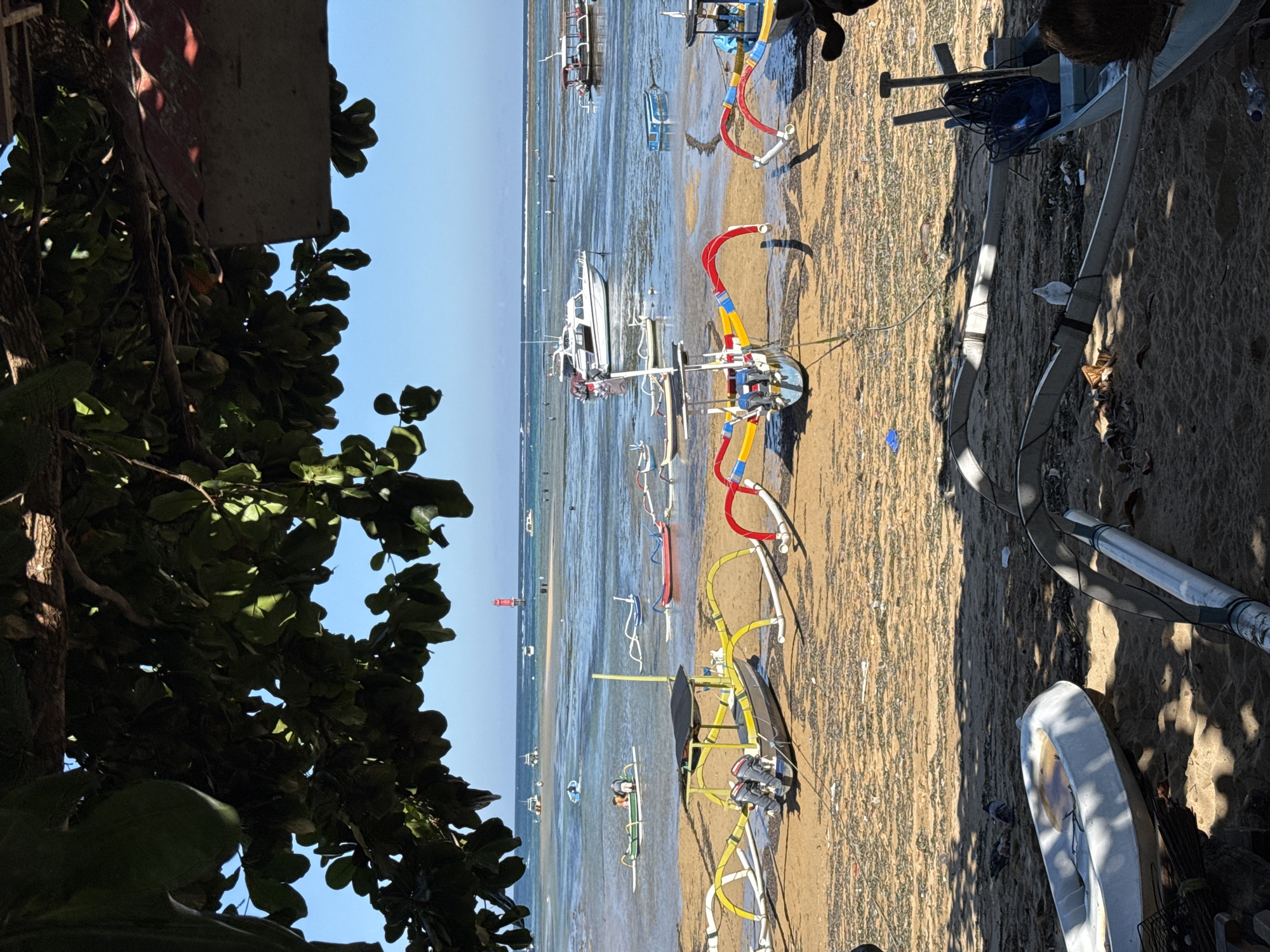 The image displays a vibrant coastal scene, likely during low tide, populated by numerous traditional Indonesian outrigger boats (Jukungs) and some larger motorboats. The perspective is from an elevated position, looking out over a sandy beach and shallow, clear blue water towards a distant horizon.
On the left, lush green foliage partially frames the view. The sun is high, casting distinct shadows and illuminating the scene brightly, suggesting daytime, possibly morning or early afternoon, under a clear blue sky.
Many boats are either beached on the exposed sandy seabed or floating in the very shallow water. Several Jukungs feature brightly colored components, notably red, yellow, and blue accents on their outriggers and hulls, creating a lively contrast with the muted tones of the sand and water. A prominent black boat with yellow outrigger supports and two outboard motors is visible in the mid-foreground. Further out, a white motorboat and various smaller vessels dot the water. In the far distance, a small red lighthouse or navigational buoy is discernible.
One individual is partially visible near the right edge of the frame, close to a boat. The overall environment suggests a working beach and coastal community. No discernible text is visible in the image.