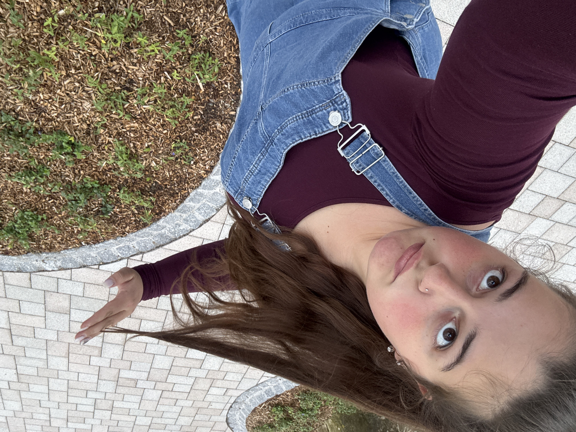 A young Caucasian female with brown hair, wearing a maroon long-sleeved shirt and denim overalls, is pictured from a high angle, looking directly into the camera. She has large brown eyes, rosy cheeks, and a nose ring. Her left hand is raised, and her hair is flowing to her right. The background consists of a paved walkway made of light-colored rectangular bricks, and an area of mulch with small green plants. The scene appears to be an outdoor public space in Schopfheim, Germany, possibly a park or a street with landscaping.