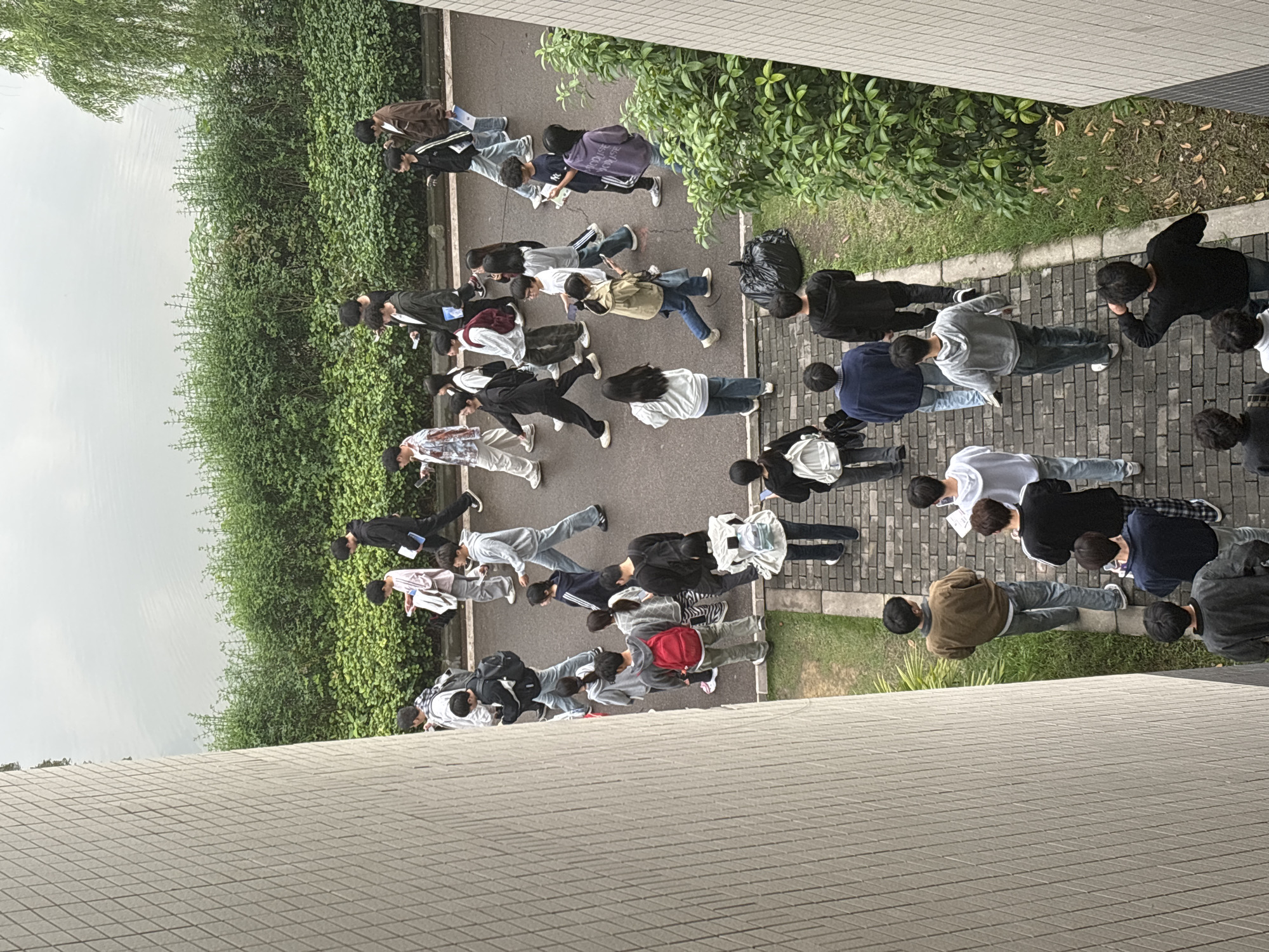 The image depicts a group of approximately 25 people walking on a paved path adjacent to a building and a green space. The path is lined with green bushes. Some individuals are carrying bags or papers, suggesting they may be students or related to an educational setting. The scene appears to be set outdoors during daylight hours, under overcast skies. The photograph was likely taken from an elevated perspective, possibly from a building overlooking the path. Jiangjin, China, is not verifiable from this image.