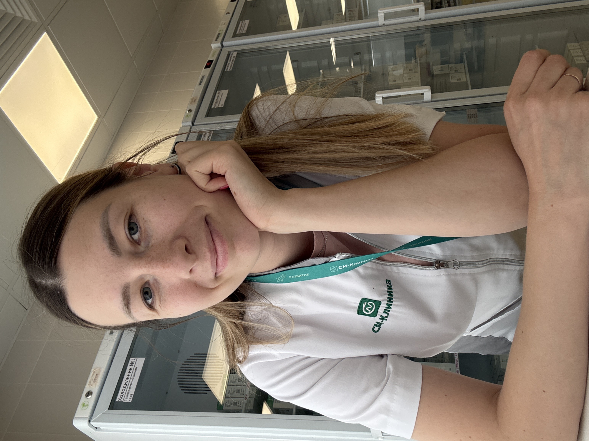 This is a selfie of a young woman, likely in her early 20s, taken indoors. She has long, blonde hair and is wearing a white lab coat over a pink top. She is leaning her head on her hand and smiling slightly at the camera. On her lab coat, there is a green lanyard with text and a logo. The text on the lanyard appears to read "СМ-Клиника" (SM-Clinic) and "РАЗВИТИЕ" (Development) and "УВАЖЕНИЕ" (Respect). Another logo on the lab coat also says "СМ-Клиника".
Behind her, there are glass-fronted cabinets or refrigerators, some of which have labels. One label clearly visible reads "Холодильник №1" (Refrigerator No. 1) and "Температурный режим +2+8" (Temperature mode +2+8). This suggests the location is a medical facility or laboratory, consistent with the lab coat. The lighting appears to be artificial, with a bright rectangular light panel visible on the ceiling in the upper left of the image. The overall impression is a candid, casual moment captured during a workday in a professional setting.