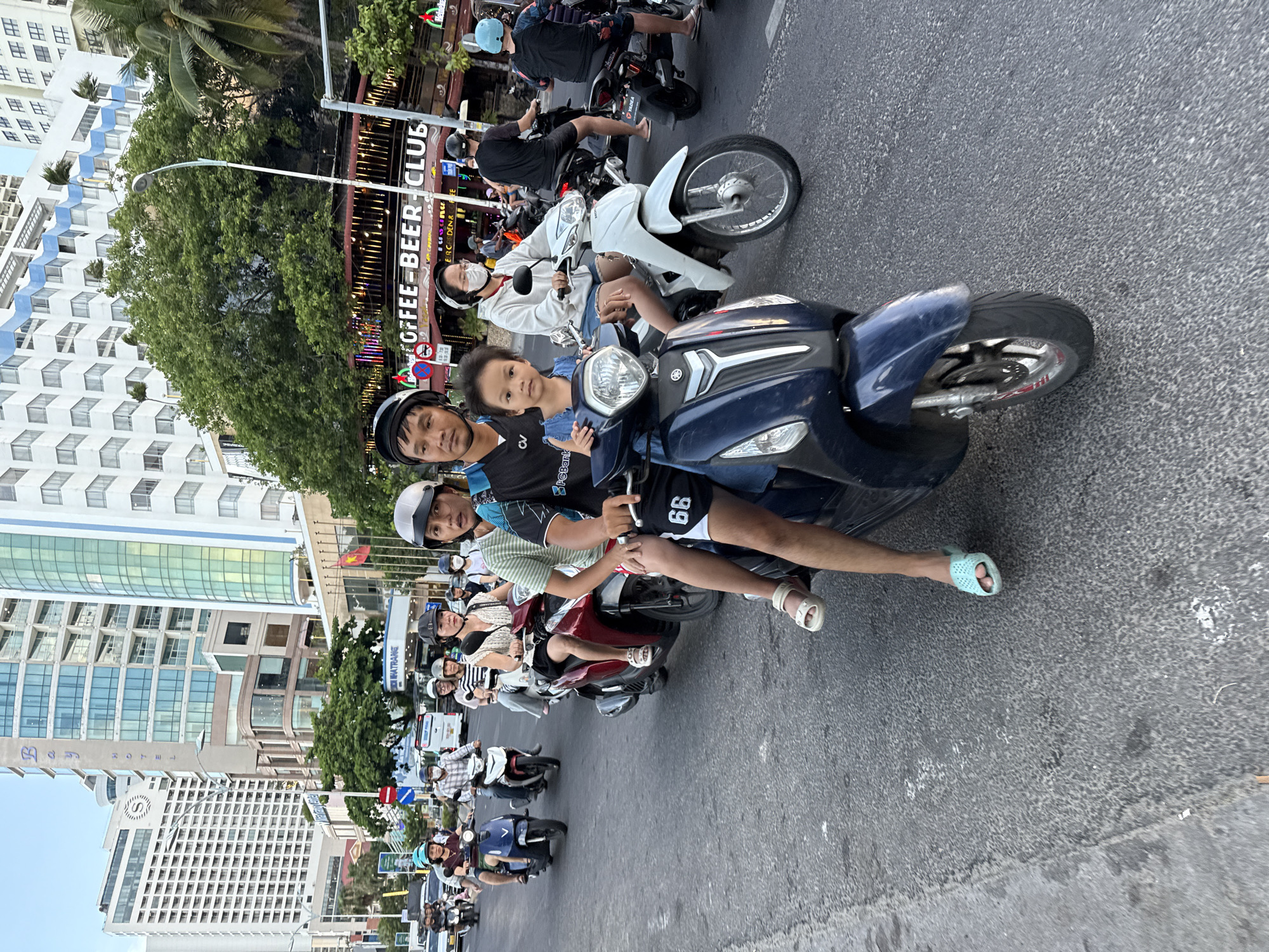 The image depicts a busy street scene in Nha Trang, Vietnam, filled with people riding motorbikes. The perspective is from one of the motorbikes, looking forward and slightly to the left. The time of day appears to be late afternoon or early evening, with the sunlight casting long shadows. The weather seems pleasant, as many people are not wearing heavy clothing.
The primary subjects are the people on the motorbikes, many of whom are wearing helmets for safety. In the foreground, a family is visible on a blue motorbike. The driver, a man wearing a helmet and a dark blue shirt with white accents, is looking towards the camera. Behind him, a child with short hair and a blue shirt sits between the driver and a woman. The woman, wearing a white top and denim shorts, is also looking forward. Another person, likely an adult, is partially visible behind the child.
The scene is set on an asphalt road, with several other motorbikes and people in the background. Buildings line the street, including a multi-story building with glass windows and a sign that reads "COFFEE-BEER CLUB". Other signs and advertisements are visible, contributing to the lively urban atmosphere. A traffic light is visible in the distance. The overall impression is one of everyday life and transportation in a bustling Vietnamese city.
Notable details include the text "CV SPGBank" and "66" on the clothing of the person in the foreground, as well as the brand "YADEA" on a motorbike in the background. The presence of "Heineken" advertising suggests a commercial area. The image captures the dynamic and often crowded nature of motorbike traffic in Vietnam.
