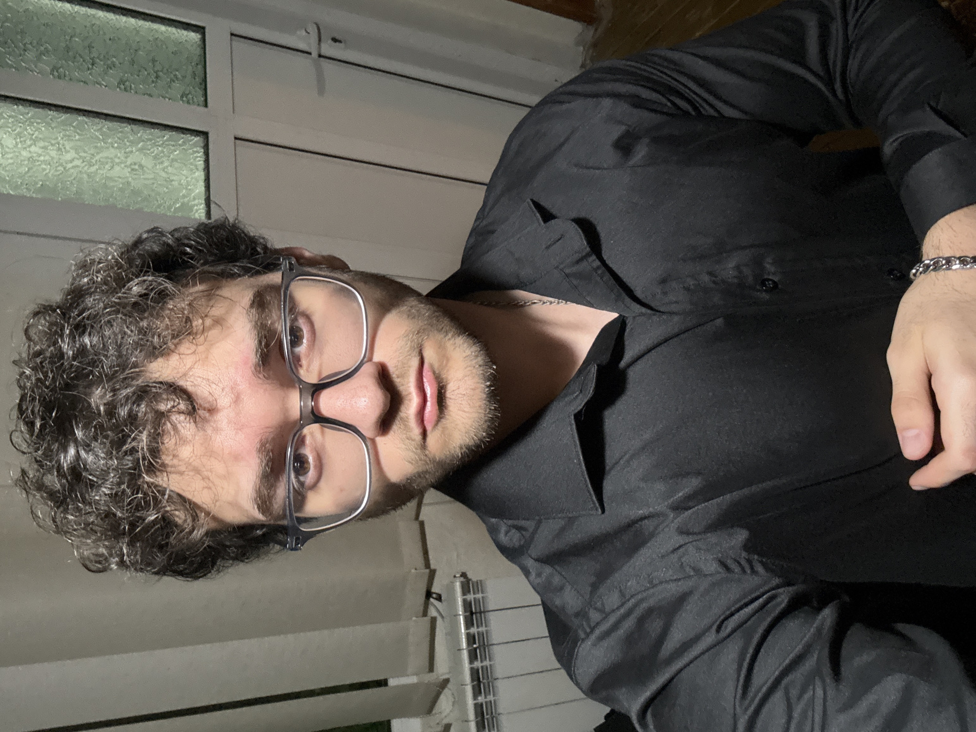 A man with curly dark hair, fair skin, and a beard is wearing glasses and a black button-down shirt. He is looking directly at the camera. He is also wearing a silver chain necklace and bracelet. The background includes a window with a patterned glass pane and a white frame. There is also a radiator. The location appears to be indoors. It is unknown if the location is in the city of Ilindentsi, Bulgaria.