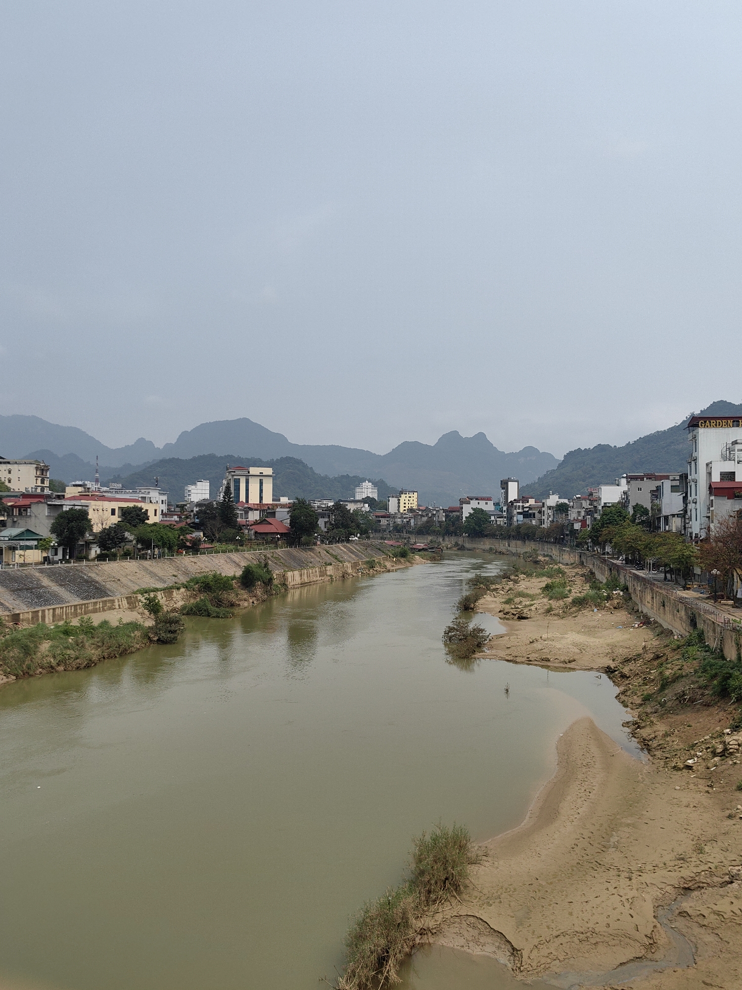 The image captures a wide-angle view of the Lô River as it flows through the city of Hà Giang, Vietnam. The river, murky with a muddy brown color, dominates the foreground. Exposed sandbanks and muddy shorelines are visible on either side of the river. The riverbanks are lined with concrete embankments and vegetation.
In the background, a cityscape is visible, featuring a mix of low-rise buildings and taller structures. The architecture appears to be a blend of residential and commercial properties. A range of mountains form a distant backdrop to the city. The sky is overcast and a dull gray.