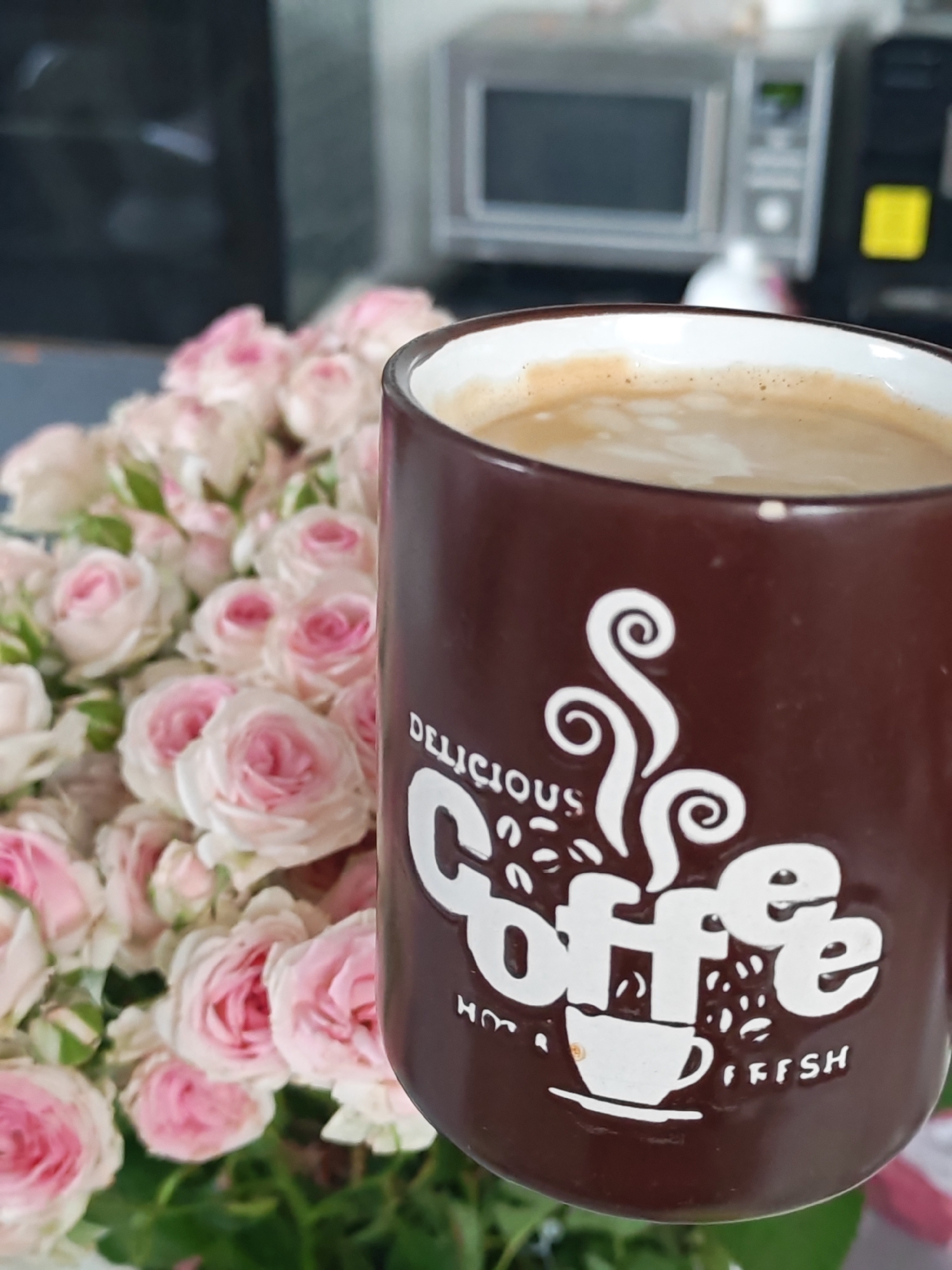 A close-up shows a brown mug with white lettering that reads "delicious coffee" and an image of a small coffee cup, positioned in front of a bouquet of pink roses. In the background, slightly out of focus, are household appliances, possibly a microwave and another appliance, suggesting an indoor kitchen or dining area. The overall impression is of a still life arrangement of coffee and flowers, with the background suggesting a domestic setting. There is no indication of the city of Rybolovo or any location information within the image.
