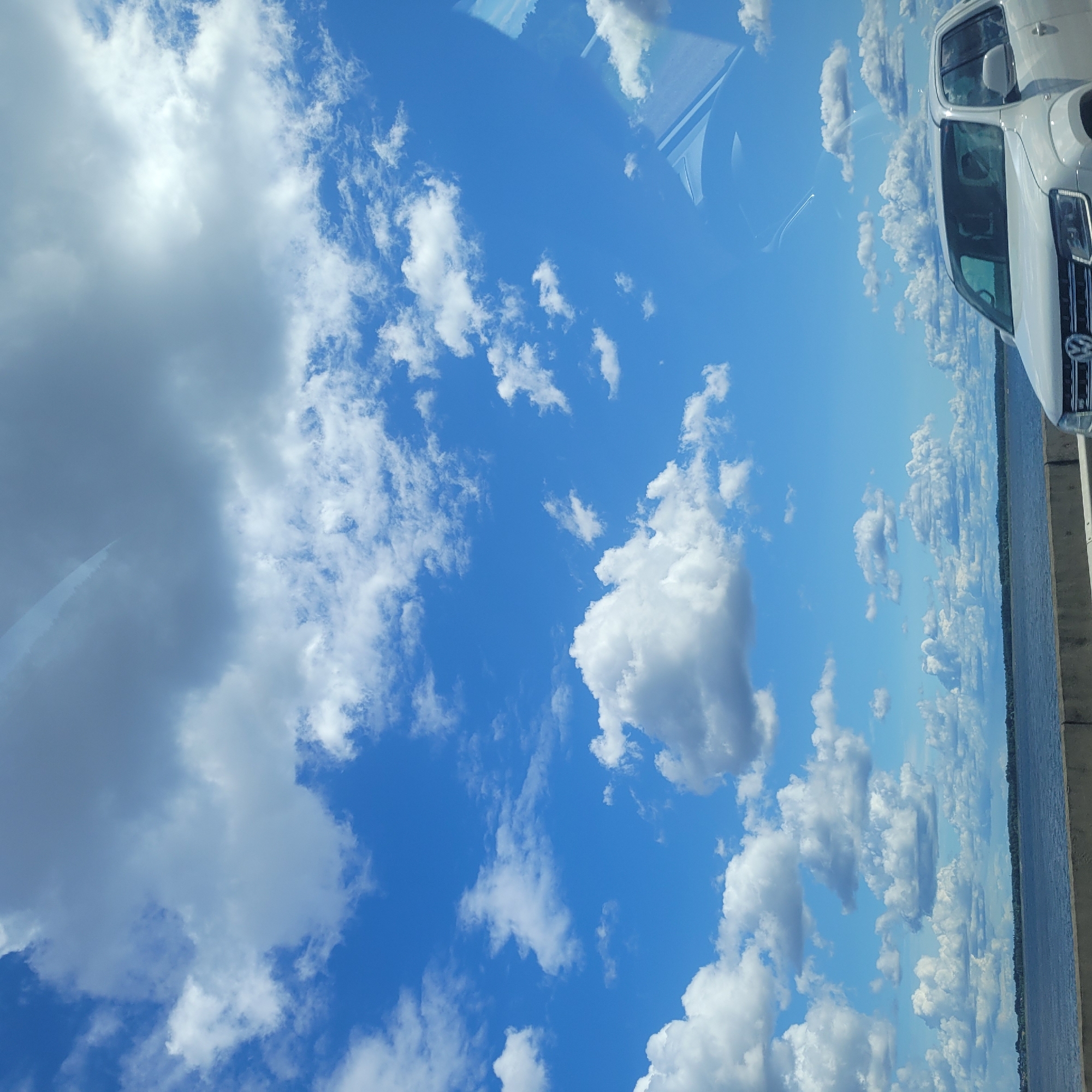The image captures a bright, sunny day with a vast blue sky filled with fluffy white clouds, some of which are large and cumulonimbus. A body of water, likely a lake or sea, stretches across the lower part of the frame, with a distant tree-lined shore visible on the horizon. In the foreground, a white pickup truck is partially visible, its front grill and headlights clearly in view. The truck appears to be on a road or highway, as suggested by the concrete barrier running along the edge of the water. The overall scene suggests a scenic drive or roadside stop with a focus on the natural landscape.