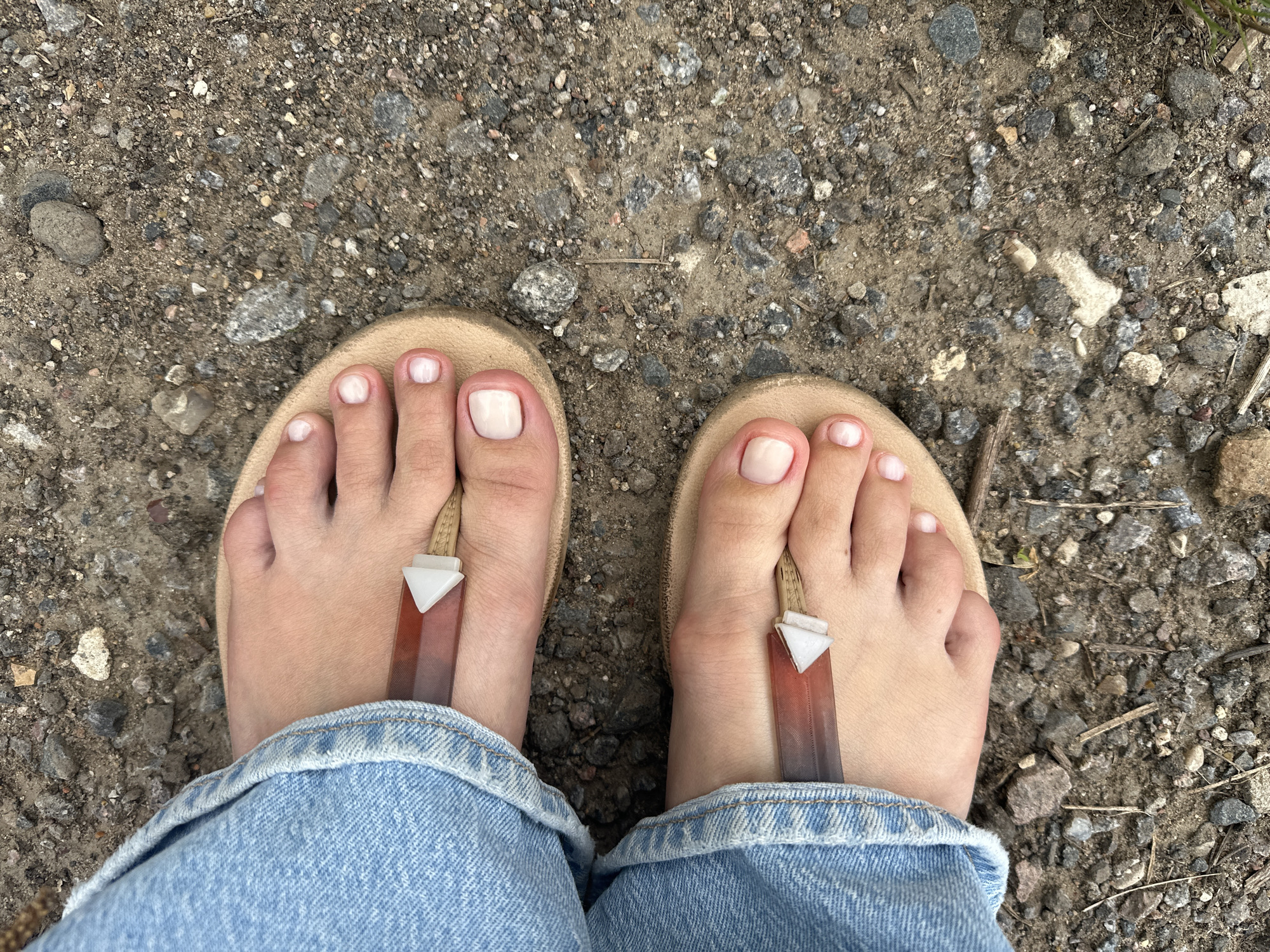 The image shows a top-down view of a person's feet wearing tan flip-flops, standing on a gravel surface. The person is also wearing light blue denim jeans that cover their ankles. Each flip-flop has a clear plastic strap with a reddish-orange gradient and a white triangular accent piece. The toes are painted with light pink nail polish. The environment appears to be outdoors, with small stones and dirt composing the ground.