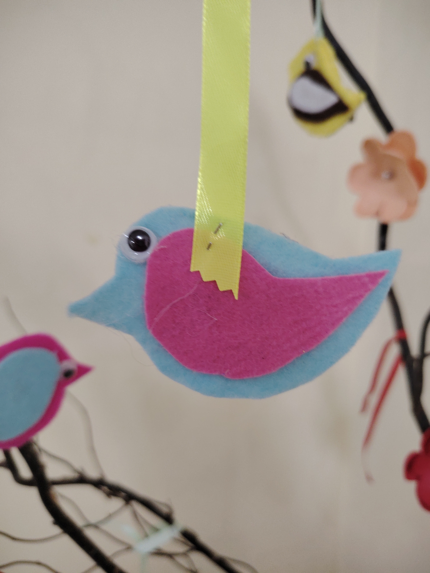 The image presents a close-up, shallow depth-of-field shot of colorful, handmade felt bird decorations, likely part of a mobile or decorative arrangement. The primary subject, in sharp focus, is a light blue felt bird with a prominent, vibrant hot pink wing and a small pink beak. A single googly eye with a black pupil gives the bird a whimsical expression. This central bird is suspended from a bright yellow ribbon, which is visibly attached to its back with two small metallic staples.
In the blurred background, other elements of the display are discernible. To the lower left, a smaller felt bird, also in blue and pink, is partially visible, appearing to perch on or near dark, bare branches that weave through the scene. On the upper right, a blurry yellow felt shape, possibly another bird or a stylized flower, hangs, alongside a soft peach-colored felt flower. The dark branches provide a natural contrast to the bright felt crafts and suggest a rustic suspension method. The background consists of a plain, light-colored surface, which is out of focus. The overall aesthetic is cheerful and crafted, possibly indicating decorations for a nursery, Easter, or a spring theme. There is no visible text in the image. The setting appears to be indoors, in Sarapul, Russia.
