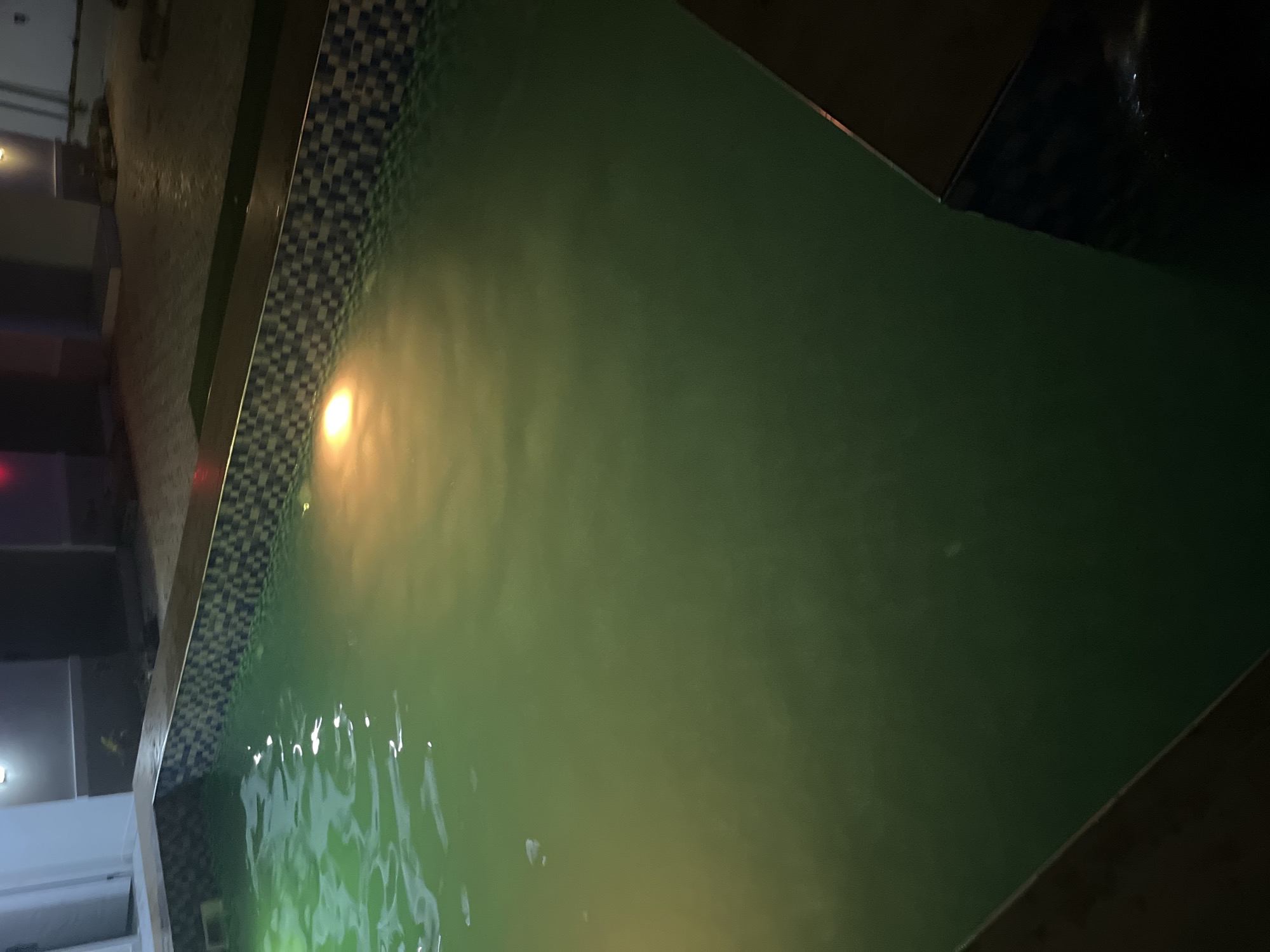 An outdoor swimming pool is pictured at night, likely in Kason, Nigeria. The water is illuminated by underwater lights, casting a greenish glow. A tiled border surrounds the pool, and in the background, a building with exterior lighting is visible. The scene suggests a residential or hospitality setting.