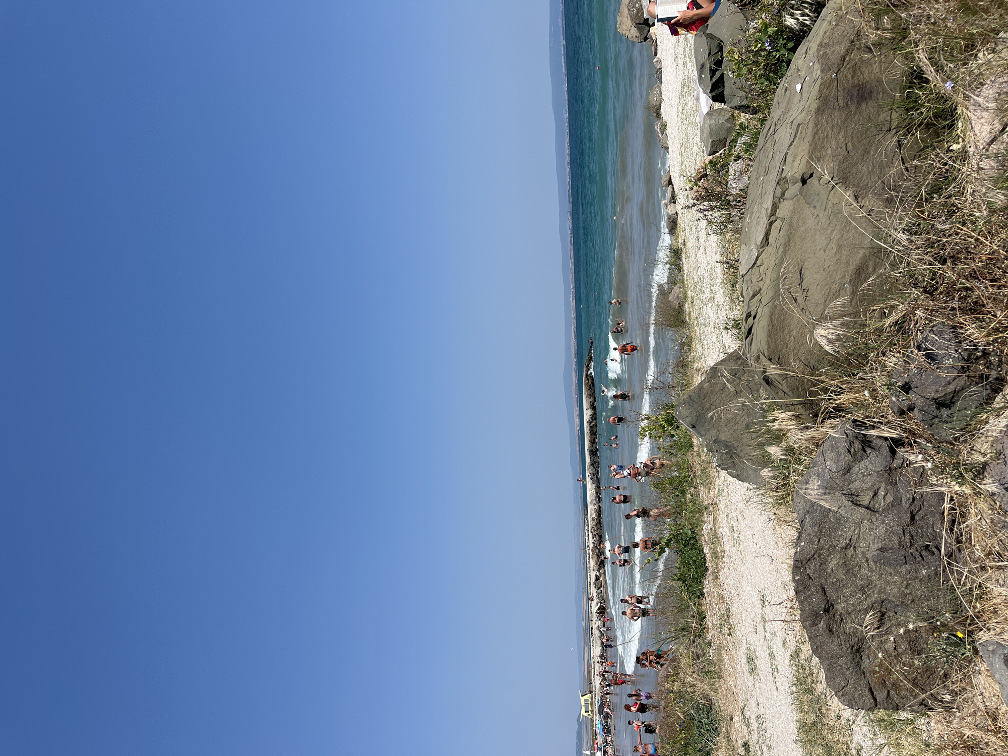 This image depicts a crowded beach scene in Pomorie, Bulgaria, under a clear blue sky. The foreground is dominated by large, rough, dark-colored rocks and dry, wispy grass. Beyond this, a strip of light-colored sand leads to the ocean. The water is a vibrant turquoise and appears to be moderately wavy, with whitecaps visible.
A long, man-made breakwater made of large stones extends into the sea from the left side of the frame, with numerous people gathered on it and around its base. Many individuals are in the water, swimming, wading, and playing in the waves. A few beach umbrellas are visible further down the beach on the left. The middle ground shows a dense concentration of people enjoying the beach and the sea.
In the far distance, a hazy coastline with low hills and some buildings is visible, suggesting the urban edge of Pomorie. The overall atmosphere is one of a busy summer day at the beach.