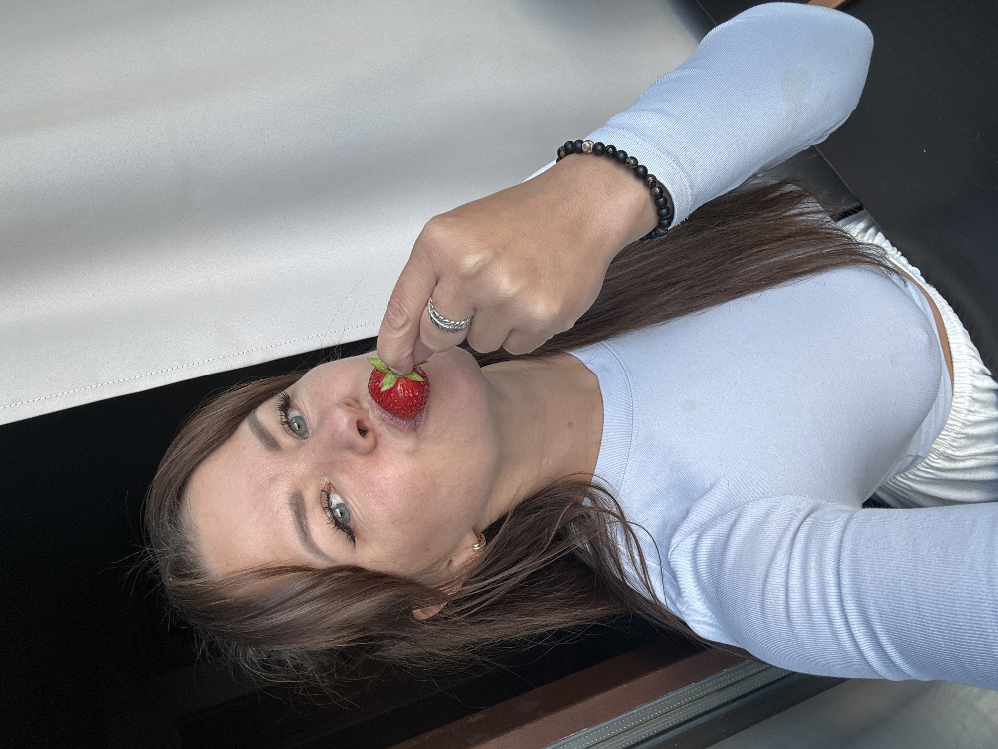 A woman with brown hair and blue eyes is holding a strawberry to her lips. She is wearing a white long-sleeved shirt and a silver ring on her right hand. There is a dark curtain on the left side of the image and a light-colored curtain on the right. The background appears to be indoors, with natural light coming from the right. The scene is a close-up shot of the woman's face and upper body.
