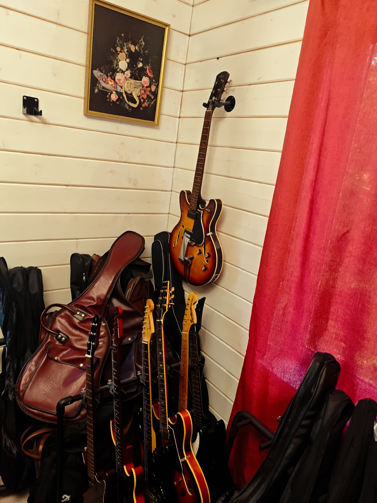 A cluttered corner of a room with light-colored, horizontal wood paneling on the walls. A framed painting of flowers hangs on the wall, above a black metal guitar wall mount (unused). An electric bass guitar hangs from another wall mount. A large, red fabric partially obscures the right side of the frame. On the floor are multiple electric guitars, some in cases, some without. A large, brown leather guitar case sits prominently among the guitar cases. The location is described as being in the city of Свистуха (кузяевский С/о), Russia, but there are no identifiable elements from the image to support that claim. No people or other actions or interactions are visible in the image.
