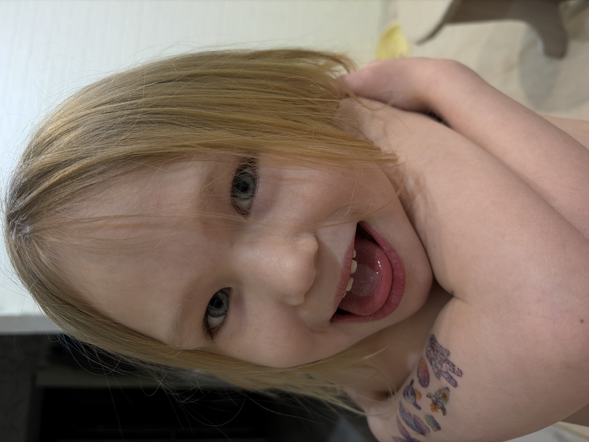 A young, light-skinned child with blonde hair and blue eyes is posing indoors. The child is smiling with their mouth open, tongue visible. On the child's left arm there are temporary tattoos of various shapes. The background is out of focus and appears to be a wall and possibly furniture. There is no indication of Noviy, Russia in the image.