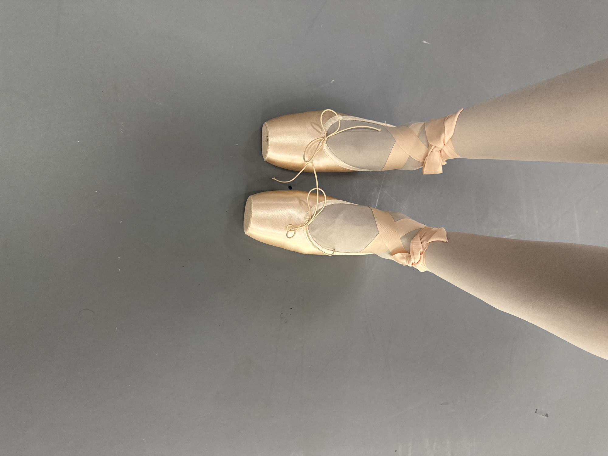 The image shows a pair of legs wearing ballet pointe shoes and tights. The shoes are a light pink color and have ribbons tied around the ankles. The tights are a similar shade. The legs are positioned against a gray background. There is no information in the image to suggest it was taken in Tbilisi, Georgia.