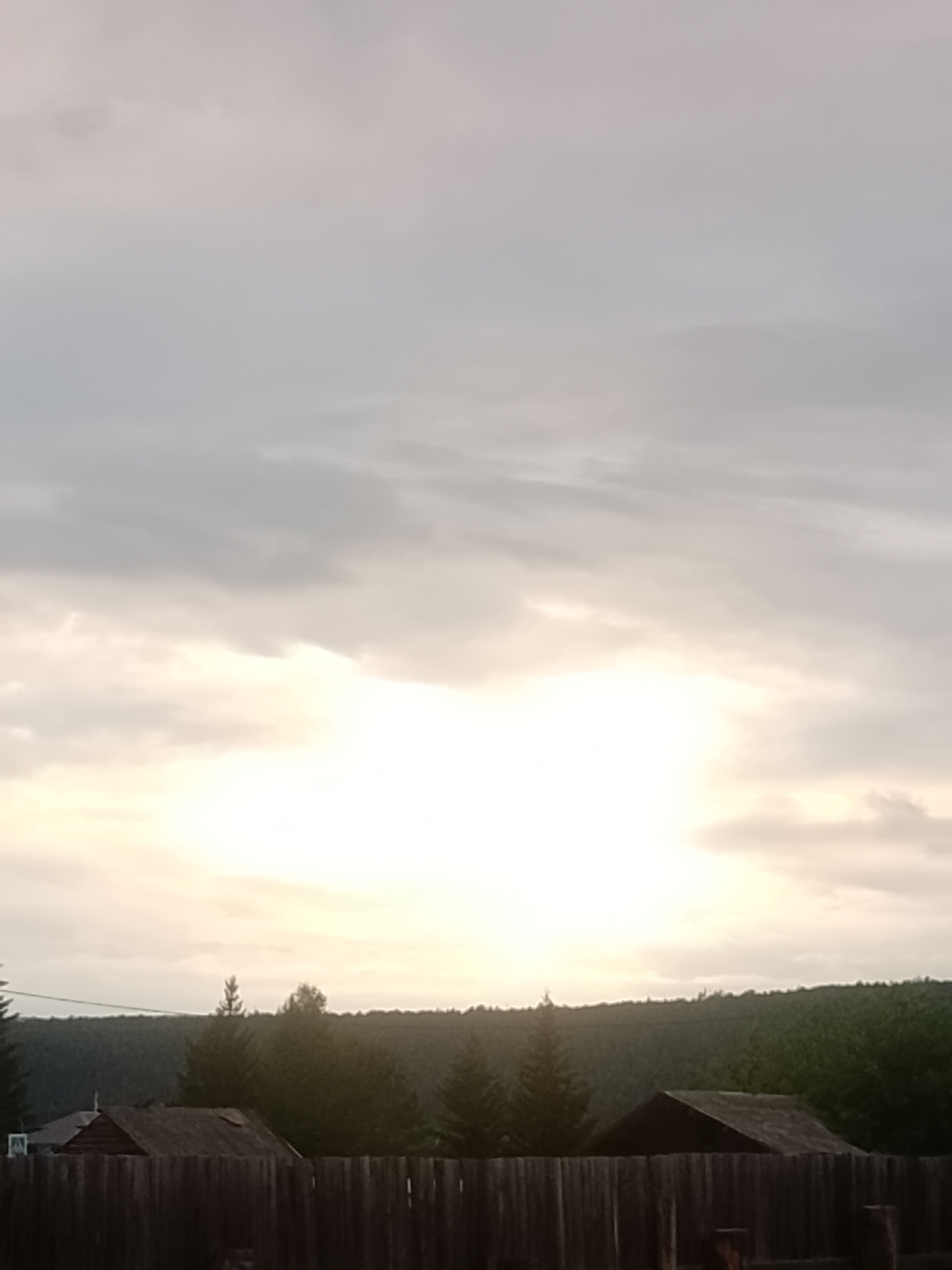 This is a vertical, low-angle, full shot of a rural scene in Chikan, Russia. The scene is dominated by a hazy, bright sky with muted clouds, suggesting either dawn or dusk. A bright patch of light in the center of the frame indicates the sun's position, either rising or setting. Below the sky, a dense line of dark green trees forms a silhouette against the horizon, with a rolling, forested hill visible beyond. In the foreground, there is a rustic wooden fence. Behind the fence, partially obscured by shadows and the fence itself, are the roofs of several small, simple buildings, likely homes or sheds, with dark wooden walls. One of the structures appears to be made of rough-hewn logs. A single, slender evergreen tree stands prominently to the left of center, in front of the denser tree line. The overall atmosphere is serene and peaceful, evoking a sense of quiet rural life. There is no visible text in the image.