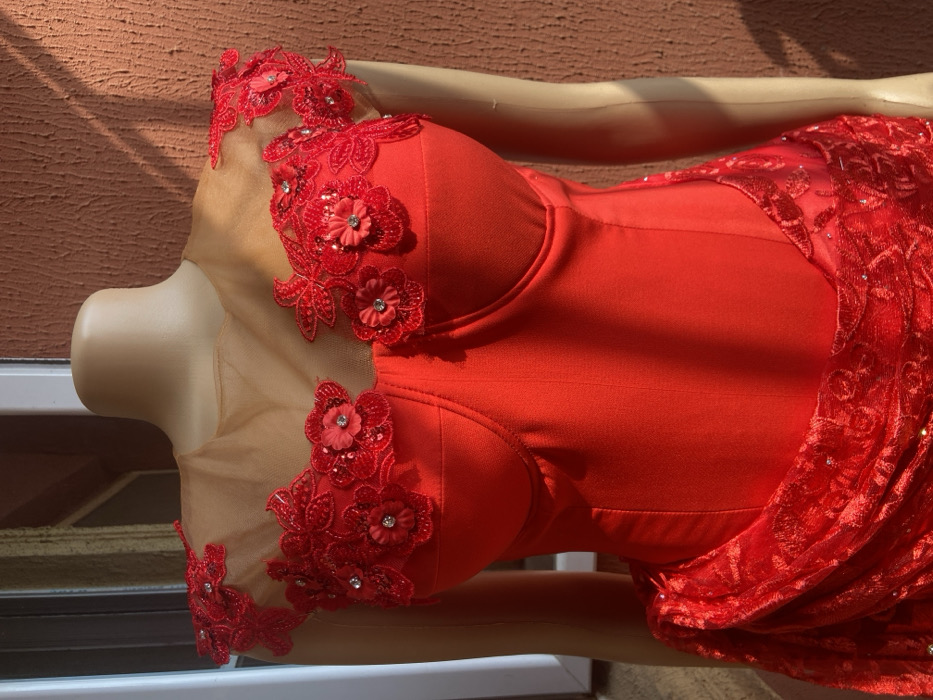 The scene depicts a red formal gown displayed on a light-skinned, armless female mannequin, set outdoors against a building in Oba-Aboji, Nigeria. The dress features a sheer, light brown illusion neckline adorned with intricate red floral lace appliques, some accented with small red fabric flowers and central clear rhinestones, extending across the shoulders and upper chest. The bodice is structured with visible bust cups and vertical seam lines, indicating a form-fitting design. The lower section of the dress, partially visible at the bottom left, consists of draped, ruched red fabric with a patterned lace overlay, which appears to contain subtle reflective elements. The mannequin and dress are positioned against a textured, light brown wall, possibly stucco or concrete. A vertical white architectural element, likely part of a door or window frame, is visible on the left. Strong natural sunlight illuminates the scene, casting distinct shadows on the wall behind the mannequin.
