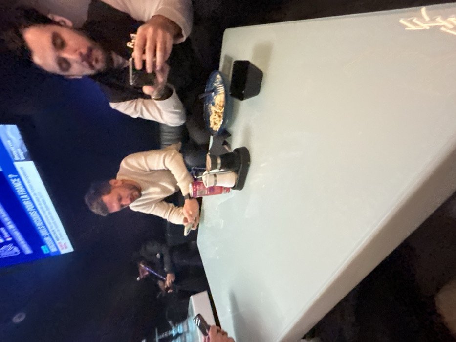 The image displays an indoor scene within a dimly lit commercial establishment, likely a lounge or cafe, situated in Illkirch-Graffenstaden, France. Four adult males are visible, primarily focused on their personal electronic devices.
In the foreground, a large, light-colored, reflective rectangular table is present. Partially visible on the left side of the table are hands holding a smartphone. Further right on the table, a red beverage can stands next to a black-based salt and pepper shaker set. Adjacent to these are a blue bowl containing partially consumed pasta with a fork, and a small, empty black square bowl.
Two males are seated at the table in the mid-ground. The male on the left, characterized by short brown hair and a beard, wears a light-colored long-sleeved top and is actively viewing a smartphone held in his hands. To his right, the second male has dark hair and a beard, wears a white long-sleeved shirt beneath a dark vest displaying "R.1" text, and is also engaged with a smartphone in his hands.
In the background, on the far left, a third male is dimly perceptible, wearing a dark cap and jacket. He holds a hookah pipe to his mouth while simultaneously looking at a smartphone. Two light-blue shisha pipes are positioned on a separate white table to his left. A fourth, unidentifiable figure is seated further back, also appearing to use a phone. A large television screen is mounted high on the dark wall, displaying French news captions, including "DOSSIER : QUELS RECOURS POUR LA FRANCE ?". The seating throughout the establishment consists of dark, upholstered couches or benches. The overall atmosphere suggests a casual social gathering where individuals are predominantly engaged with their smartphones.