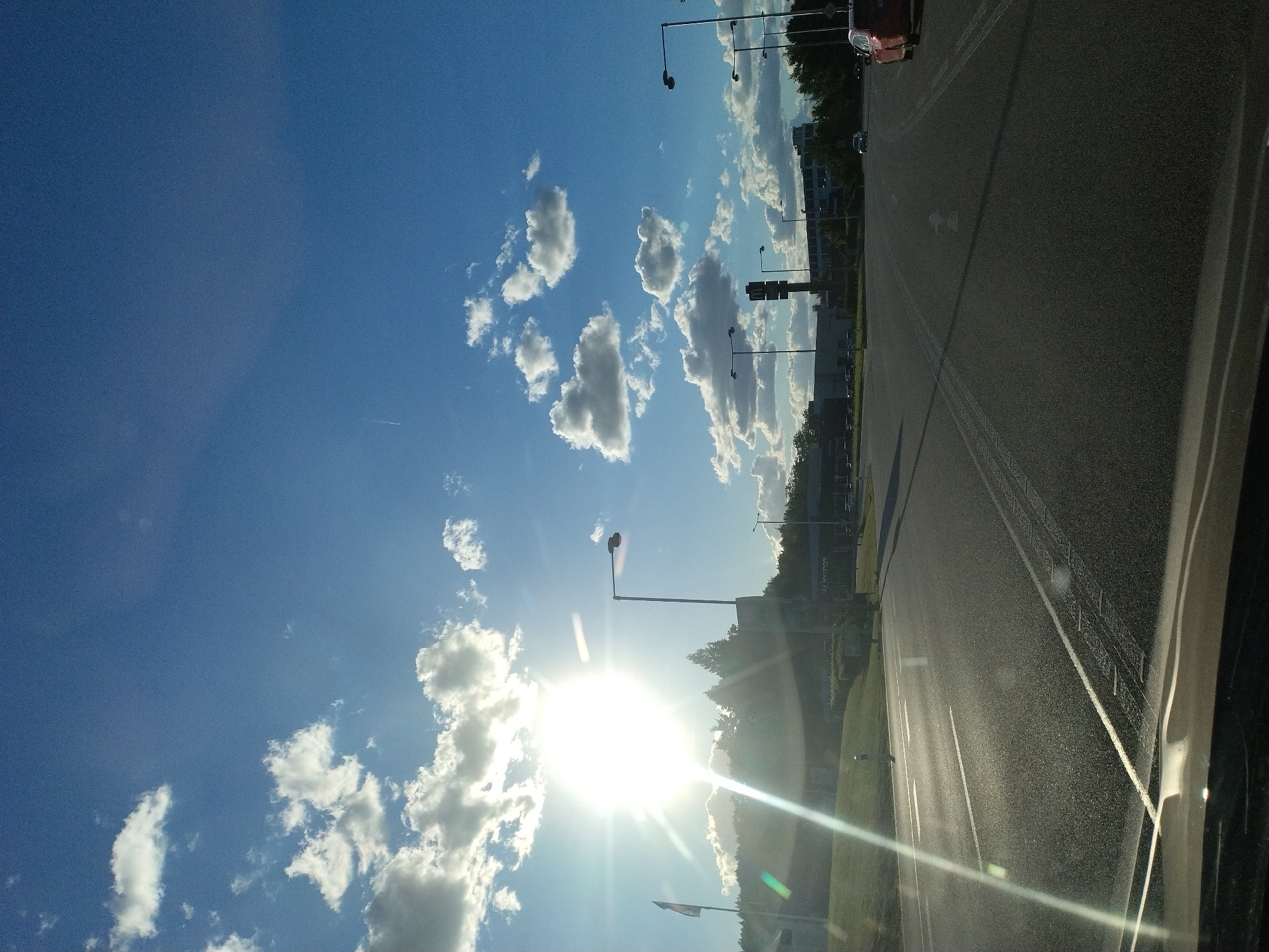 The image shows a sun-drenched highway or road with a bright sun and scattered clouds in the blue sky. The perspective is from inside a vehicle, looking out. In the distance, there are buildings, trees, and streetlights. One street light on the left has a red light at the top. A red car is visible on the right side of the road. The sun's glare is intense, creating lens flares across the image. The road has lane markings. On the left side, there is a grassy embankment with a flag pole. In the middle ground, there's a large sign with a prominent "M" logo, likely for McDonald's, and a Hyundai dealership sign in the background. The overall scene suggests a commercial or suburban area on a clear day.