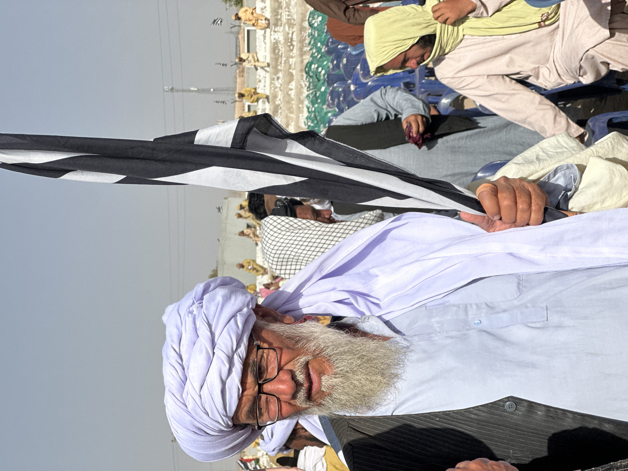 The image captures an outdoor gathering under a clear, bright daylight sky in what appears to be an arid region, consistent with Pishin, Pakistan.
In the immediate foreground, an older man with a long, prominent grey beard and glasses is the central subject. He wears a light blue shirt and a dark vest, topped with a white turban (pagri). He looks towards the viewer with a calm, slightly neutral expression.
A black and white flag is held upright to the left of the older man, obscuring part of his body. A hand with a bandaged thumb is visible gripping the flag's pole. Behind the flag, several other men are present. One man, partially visible, wears a light yellow head covering and light-colored clothing, seated on a blue plastic chair. More blue plastic chairs and large blue water containers are visible in the mid-ground.
Further in the background, indistinct figures of people, some wearing traditional attire, are visible against a backdrop of light-colored ground or structures and distant utility poles. The scene suggests a public assembly, possibly a community meeting, religious gathering, or political rally. The clothing worn by the attendees, including turbans and traditional garments, aligns with cultural styles found in the region. There is no decipherable text visible on the flag or elsewhere in the image.