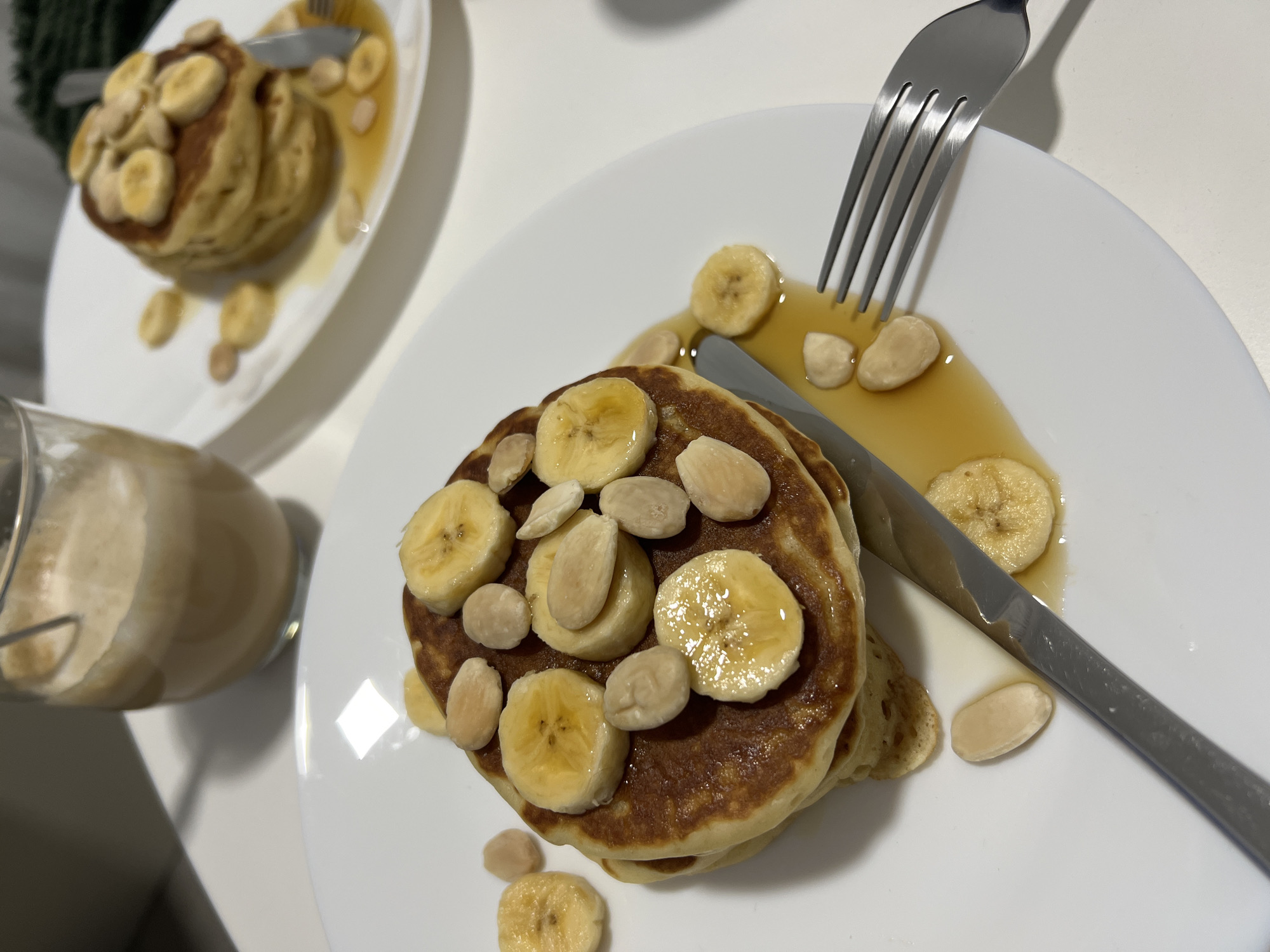 The image depicts two servings of pancakes topped with sliced bananas and almonds. Each serving is presented on a white plate, accompanied by a fork and a knife. A light-colored liquid, possibly syrup, is drizzled around the pancakes. To the left of the frame, a glass jar containing a creamy beverage is visible. The scene is set indoors on a white surface. The location is not discernible from the image.