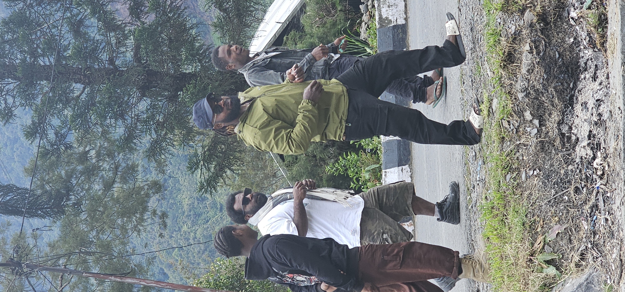 In the city of Puncak Jaya, Indonesia, four individuals walk along a paved road. The person in the foreground on the right wears black pants and white slides. To their left, a person in a green jacket and black pants walks with a blue cap and sunglasses. Next, a person in a white t-shirt and camouflage shorts walks with sunglasses and a scarf. The person on the far left wears a black hoodie and brown pants. The background features lush green hills, trees, and a cloudy sky, suggesting a mountainous or elevated terrain. The road has a white and gray striped curb, and the ground beside it is rough and covered with grass, dirt, and debris.