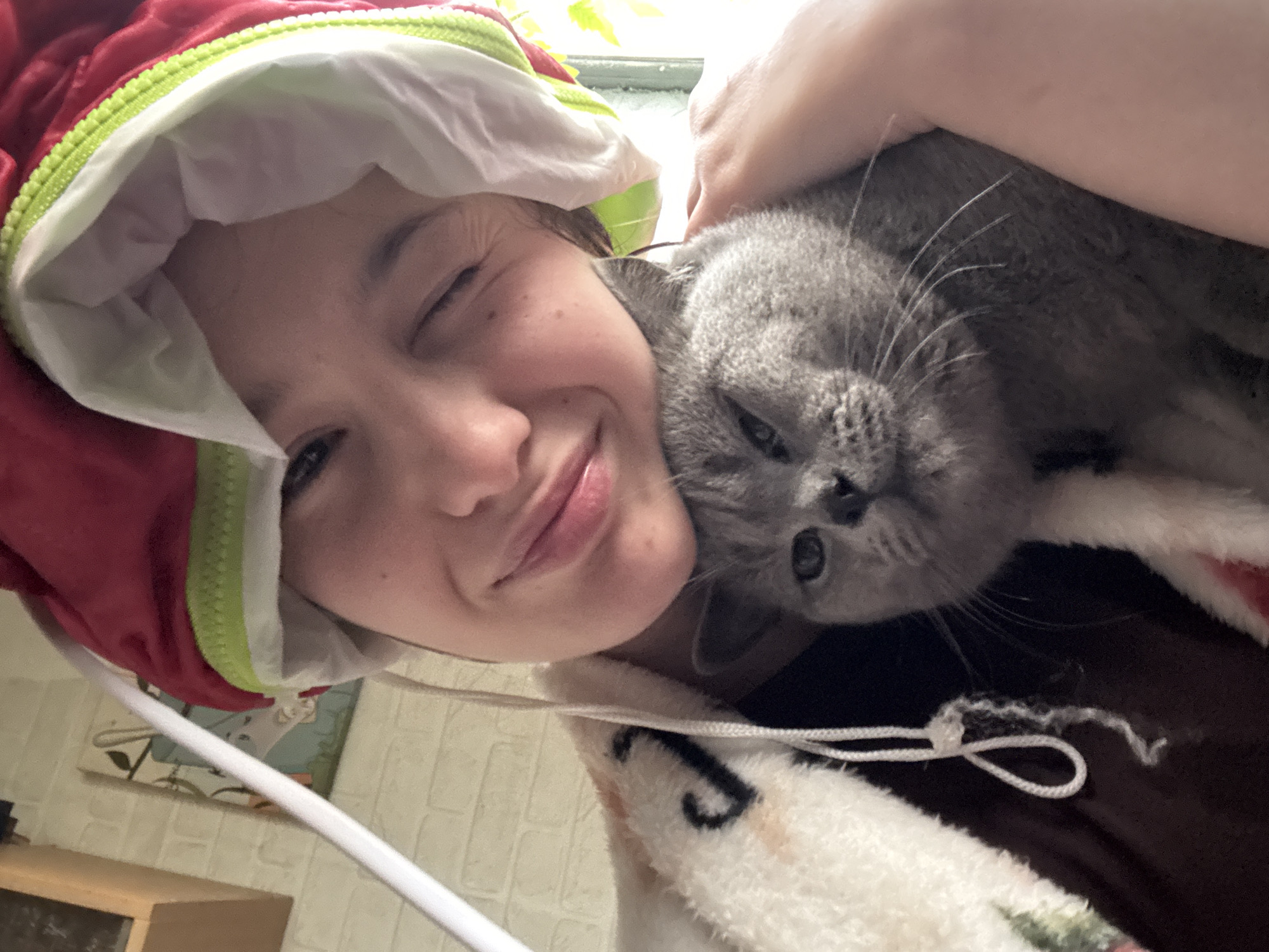 A woman with a red and white padded hood smiles and winks, a gray cat snuggled against her face. The location cannot be determined with the provided information.
