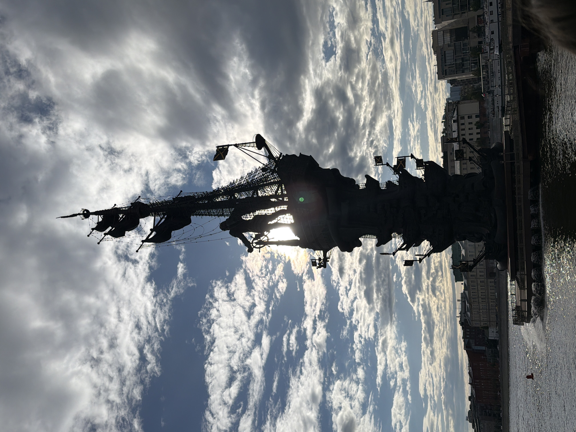 The image shows the Peter the Great Statue in Moscow, Russia, silhouetted against a sky with dramatic clouds. The statue depicts a ship, with Peter the Great at the helm. The Moskva River is visible in the foreground, with buildings along the embankment.