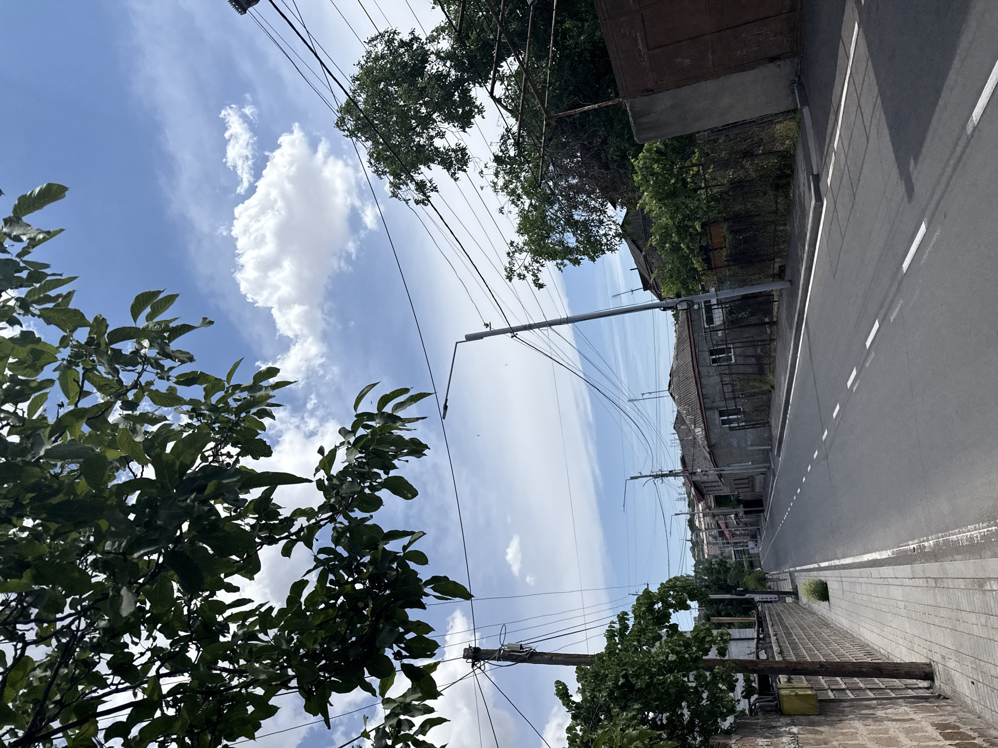 A downward-angle, eye-level shot captures a street scene in a city. The road, which is paved and has white lane markings, stretches into the distance. On the left side of the road, there is a sidewalk with brick walls bordering it, and a wooden utility pole stands nearby. Trees with green foliage frame the top and left sides of the image, with some branches hanging over the road. The right side of the road features houses with gray roofs and walls, some with fences and gates. A brown metal gate is visible in the background on the right. The sky is blue with scattered white clouds. Power lines are strung across the scene, supported by utility poles. The overall impression is a quiet street in a residential area.