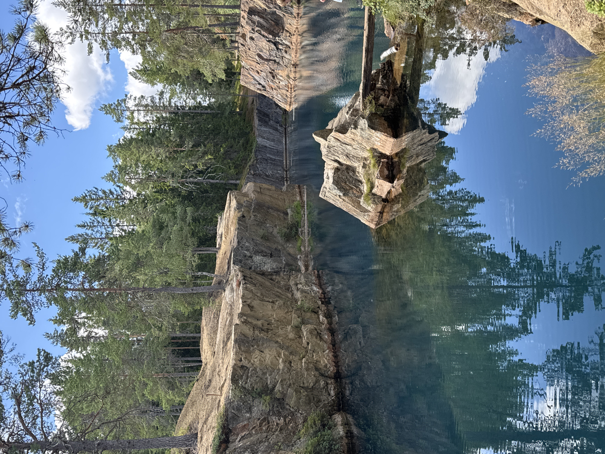 The image depicts a quarry lake located in Säter, Sweden. The water is a vibrant, clear blue, reflecting the surrounding evergreen forest and the sky above. Rocky cliffs, characteristic of a quarry, rise from the water's edge. A large rock formation sits in the middle of the lake, with a fallen log serving as a makeshift bridge to a smaller rock outcrop. A person, partially visible on the right side of the image, is standing near the edge of the quarry, looking out at the lake. The scene is bathed in natural sunlight, suggesting a clear day.