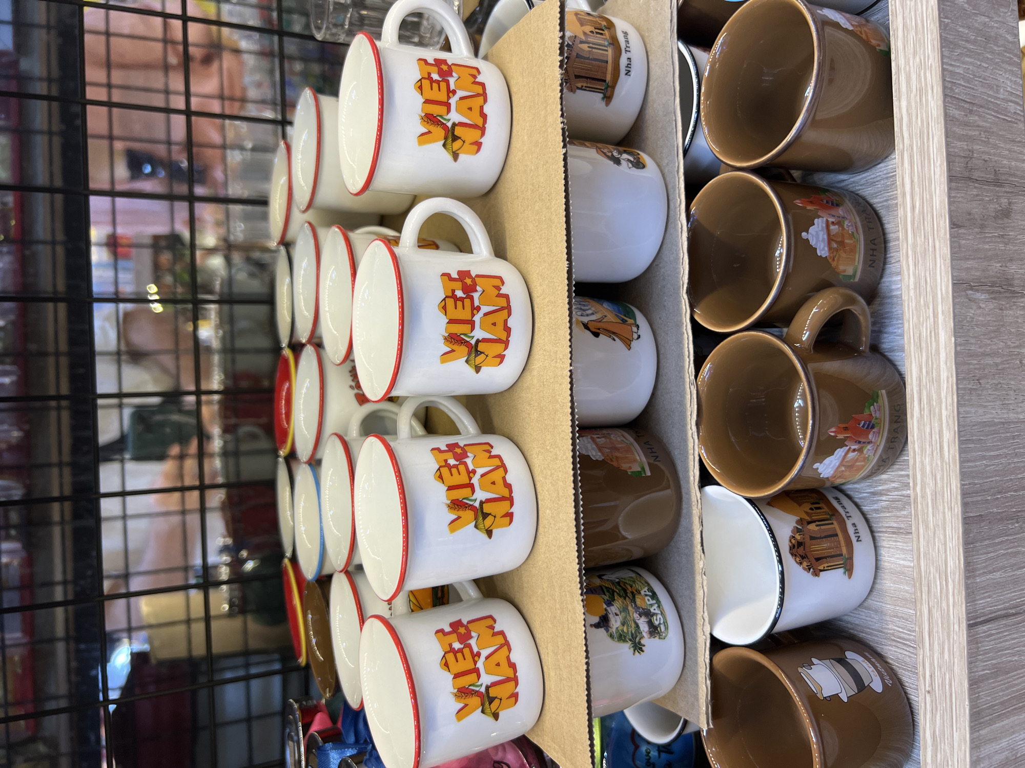 The image displays a collection of souvenir mugs arranged on shelves in a retail setting. The mugs are primarily white with red rims, featuring the text "VIET NAM" in orange lettering, along with an image of a conical hat and a Vietnamese flag. Some mugs have additional designs, such as landscapes or depictions of Vietnamese culture, with some explicitly labeled "NHA TRANG," indicating their origin or theme related to the city of Nha Trang, Vietnam. The mugs are stacked and displayed in a manner consistent with a shop or market stall. The background suggests an indoor environment with shelving and other products, likely a souvenir shop.