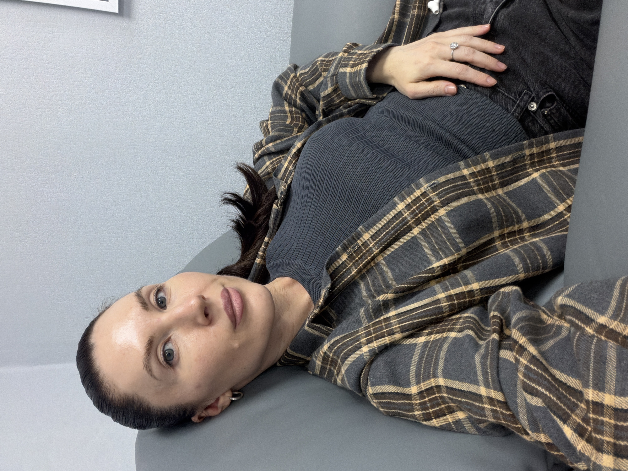 A woman lies on a gray surface. She wears a gray ribbed sweater and a gray and tan plaid shirt. Her hair is dark, and she is wearing a ring on her left hand. She is looking towards the camera. The background is a gray wall. The location is unknown and is not suggested by the image. There is no clear basis to indicate that the location is in Perm, Russia.