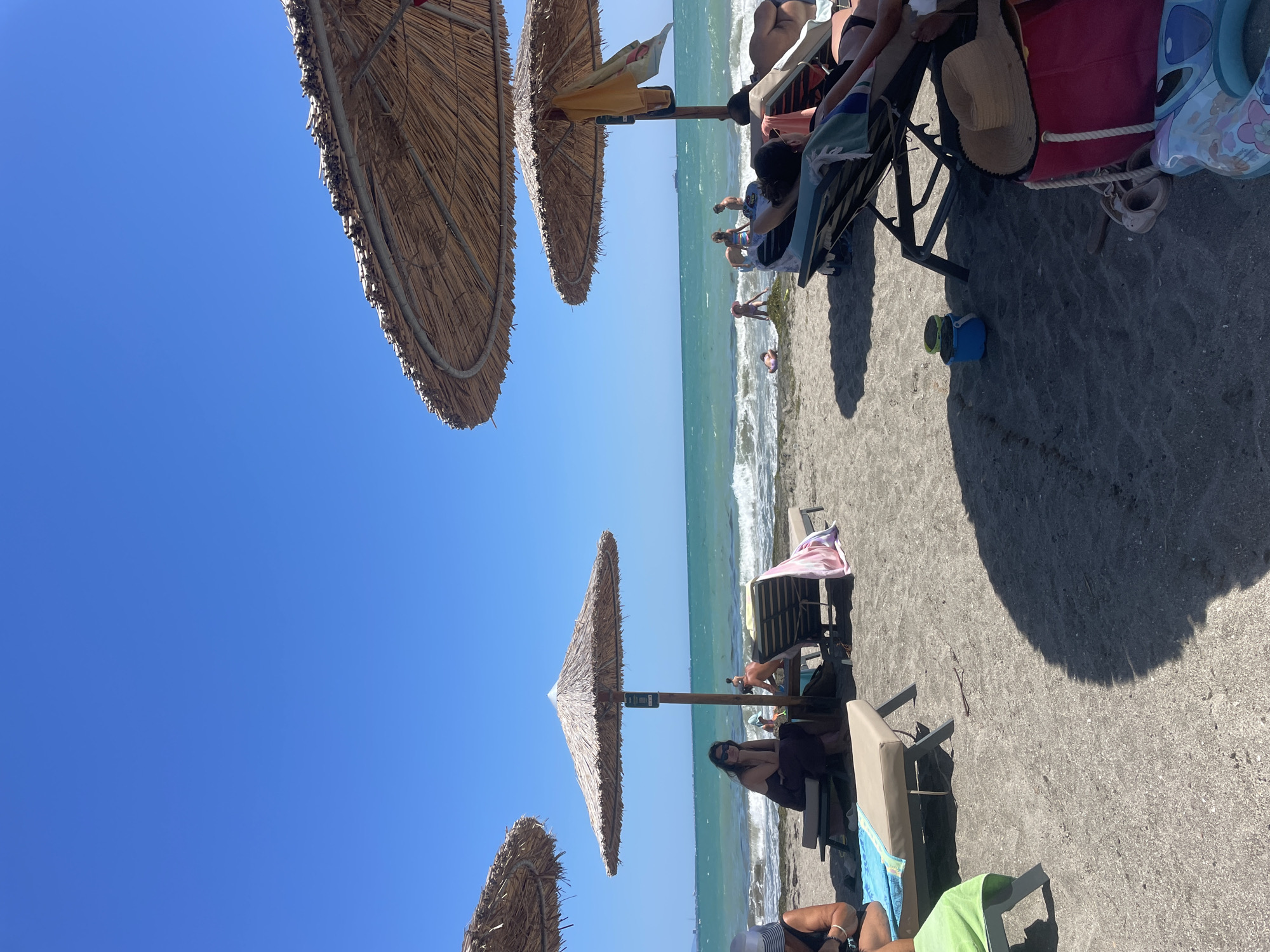 The image depicts a sunny day at a beach in Burgas, Bulgaria. The foreground is dominated by dark sand with several beach chairs and thatched umbrellas providing shade. A few people are visible, some reclining on the chairs, while others are nearer the water. The sea is a vibrant turquoise, with gentle waves rolling onto the shore. In the background, the sky is a clear, bright blue. Scattered beach items like towels, a bucket, and a beach bag are also present.