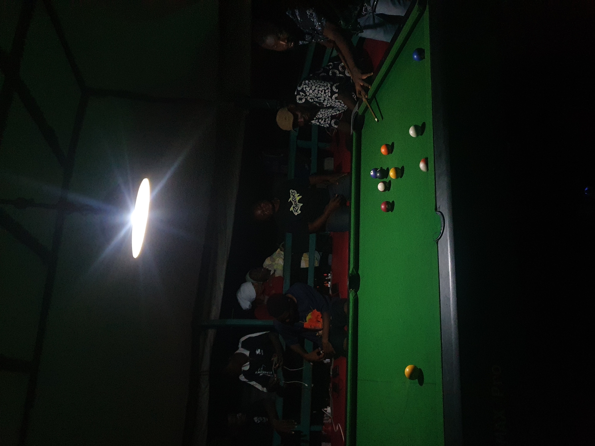 A group of men are playing pool at night in Lokoja, Nigeria. The scene appears to be an outdoor or semi-outdoor establishment, possibly a bar or recreational center, with a roof overhead and a visible bright light source illuminating the pool table. The pool table is green with several colored balls scattered across it, and a cue stick is in use by one of the players. There are at least six men visible, some actively playing and others observing. The lighting is dim, creating strong shadows. The overall impression is a casual gathering for a game of pool.