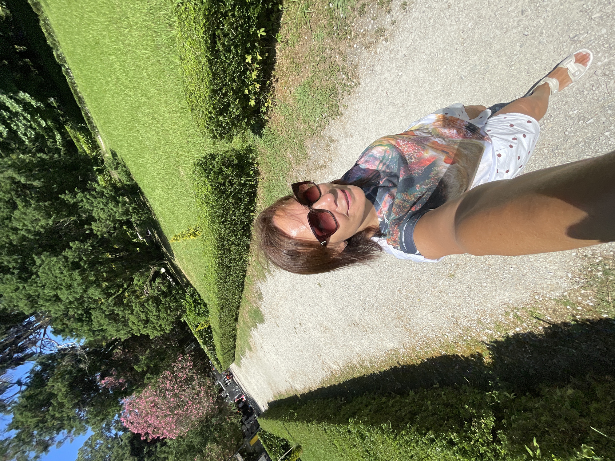 A woman with brown hair and sunglasses is taking a selfie in a park on a sunny day. She is wearing a white t-shirt with a colorful, abstract print and white shorts with brown polka dots. She is also wearing white sandals. The park has a gravel path lined with neatly trimmed green hedges. Beyond the hedges, there is a large expanse of green grass, and further in the background, there are tall trees, one of which has pink flowers. The location is identified as the city of Sirius, Russia.