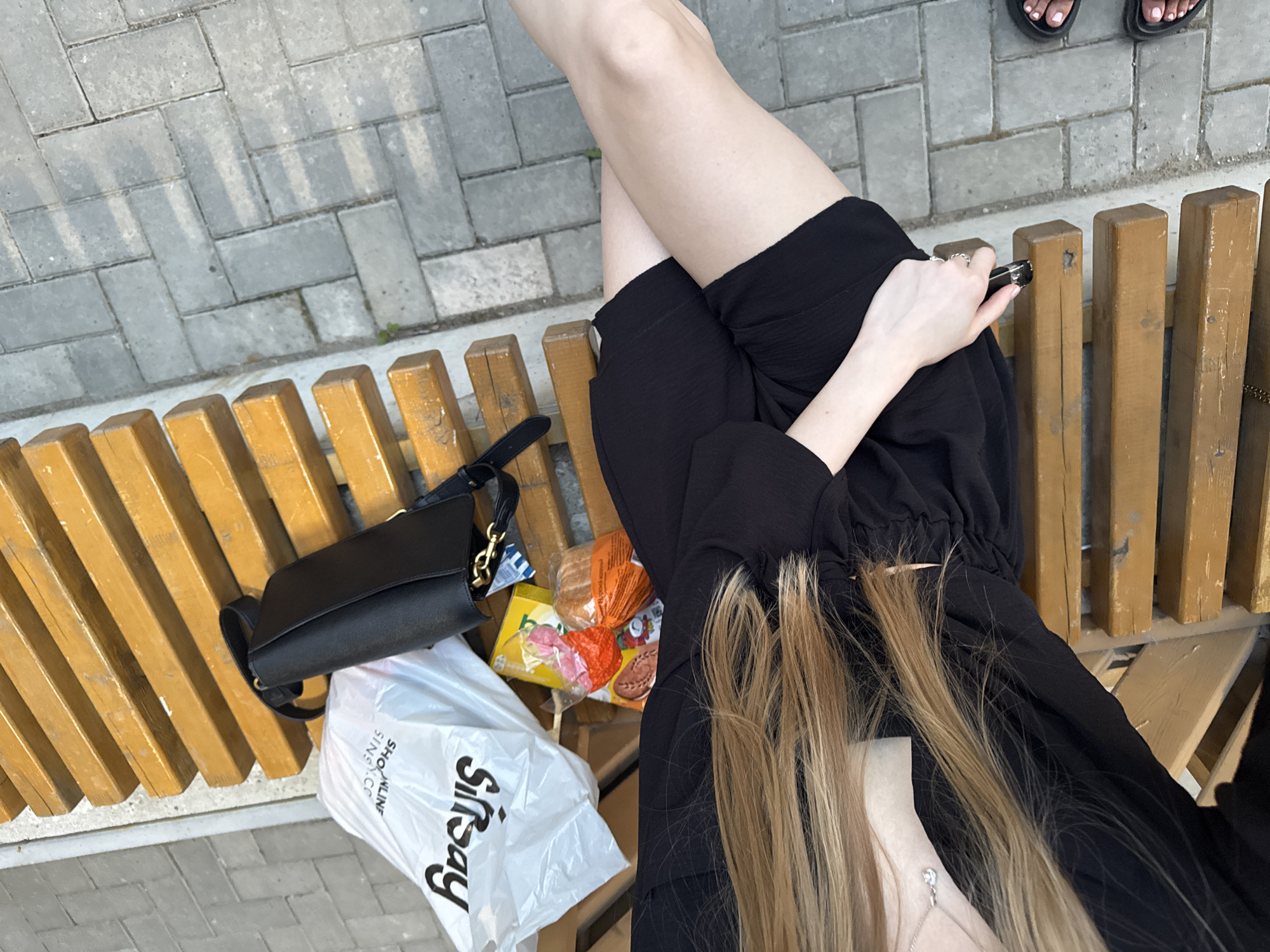 A high-angle shot depicts a woman sitting on a wooden bench in what is likely a public space. The location is possibly in Perm, Russia, as suggested by the "Sinsay" branded shopping bag visible on the bench. The woman is wearing a black dress and has blonde hair that cascades down her chest. She holds a vaping device in her hand. Adjacent to her is a black purse, a shopping bag, and what appears to be packaged food items. The bench is situated near a paved area with gray brick-like stones.