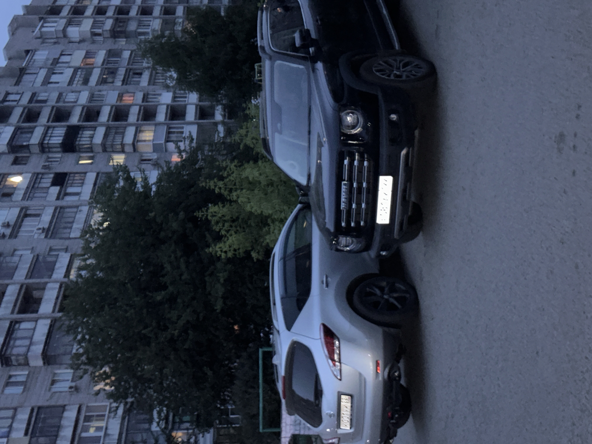 In Novoaltaysk, Russia, two cars are parked in front of a tall, multi-story apartment building. The building has numerous windows and balconies, with some lights visible inside, suggesting it is evening. Trees with dark green foliage are situated between the building and the parking area.
The vehicles are positioned close together, with a dark-colored SUV in the foreground and a silver crossover SUV behind it. The silver SUV has its rear facing the camera, and its license plate is visible, displaying the text "091 KM 82". The dark SUV is oriented with its front towards the camera, and its license plate is also partially visible. The overall scene is illuminated by ambient light, indicating it is likely dusk or early evening. The ground appears to be asphalt or concrete. There are no people visible in the frame. The image is taken from a slightly elevated perspective.
