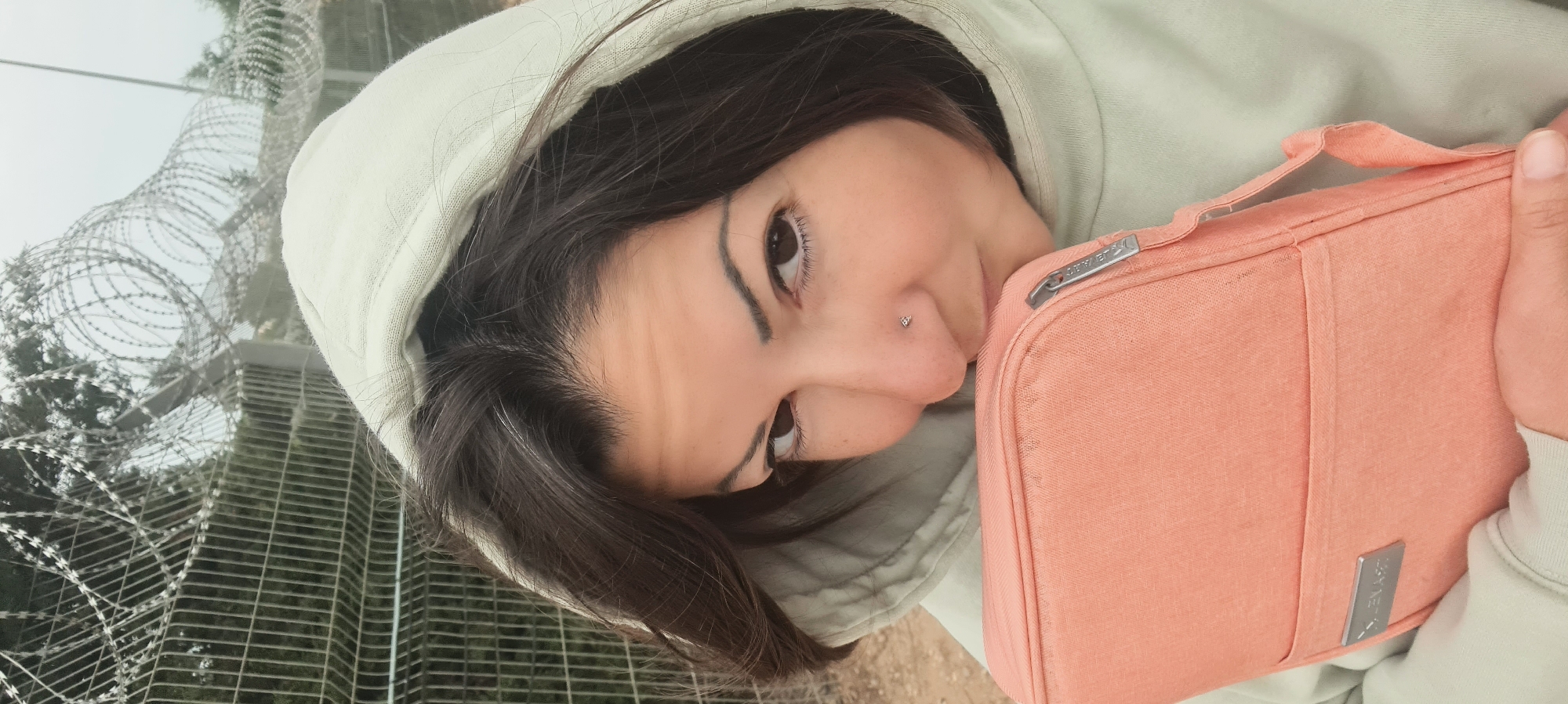 A woman in a light-colored hooded sweatshirt, holding a peach-colored bag with a silver logo in front of her, stands against a backdrop of a high security fence topped with barbed wire. The location is in the city of Lachish Regional Council, Israel, based on the barrier and implied security protocols. The sky appears overcast. The woman is angled with her head tilted, creating a selfie-like perspective.