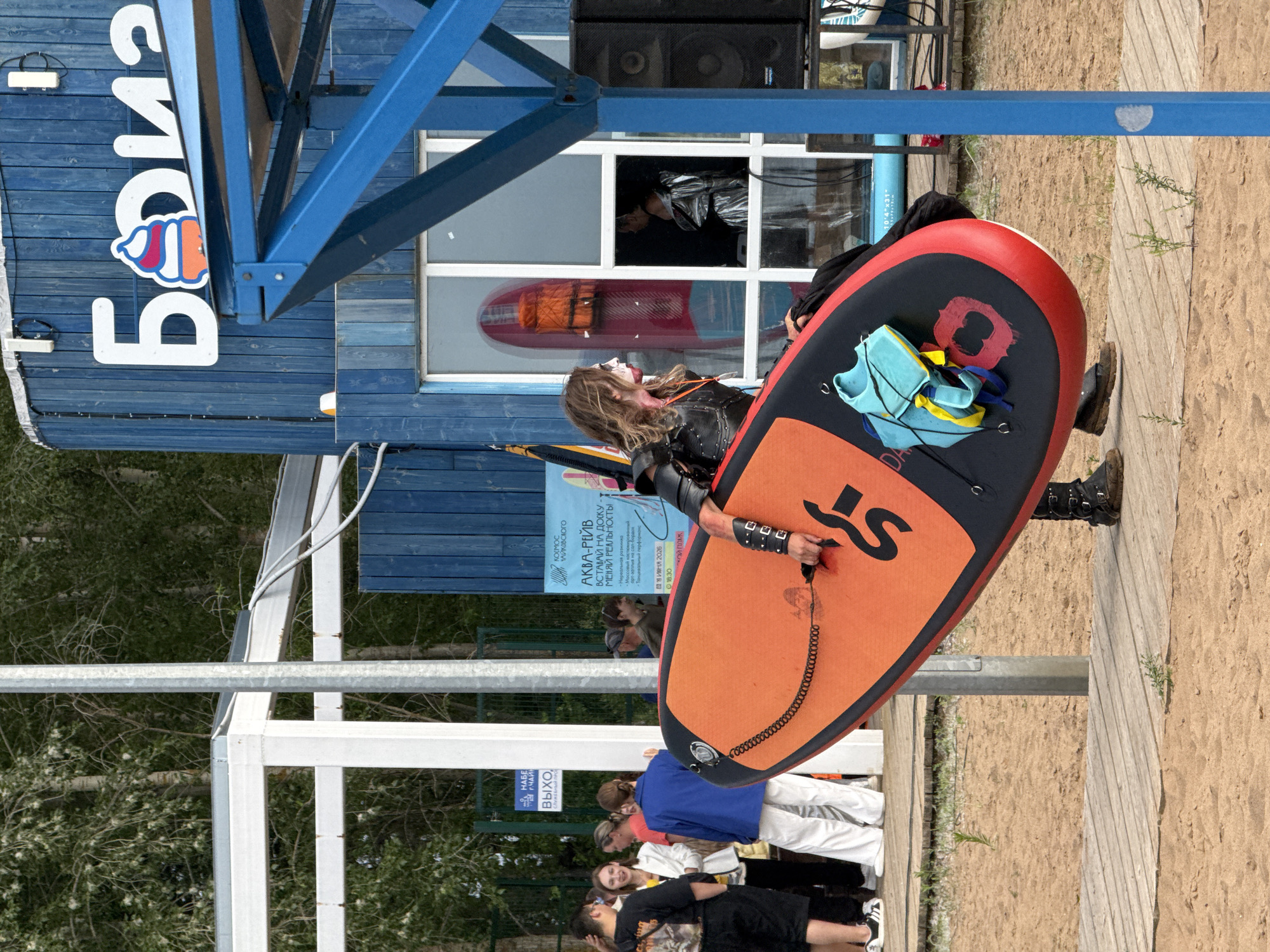 A person with long, wavy hair, wearing heavy makeup including red lipstick and dark eyeliner, and dressed in a black leather outfit with studded accessories, is carrying a bright orange and black stand-up paddleboard on a sandy beach. The paddleboard has a blue and yellow life vest attached to it. Behind the person is a blue wooden building with a sign featuring an ice cream cone logo and Cyrillic lettering that reads "Борис". A white canopy structure is visible to the left of the building, and a group of people are standing nearby. The scene appears to be set in the city of Chaikovsky, Russia, as indicated by the presence of the building and its signage, and likely a public beach or recreational area.