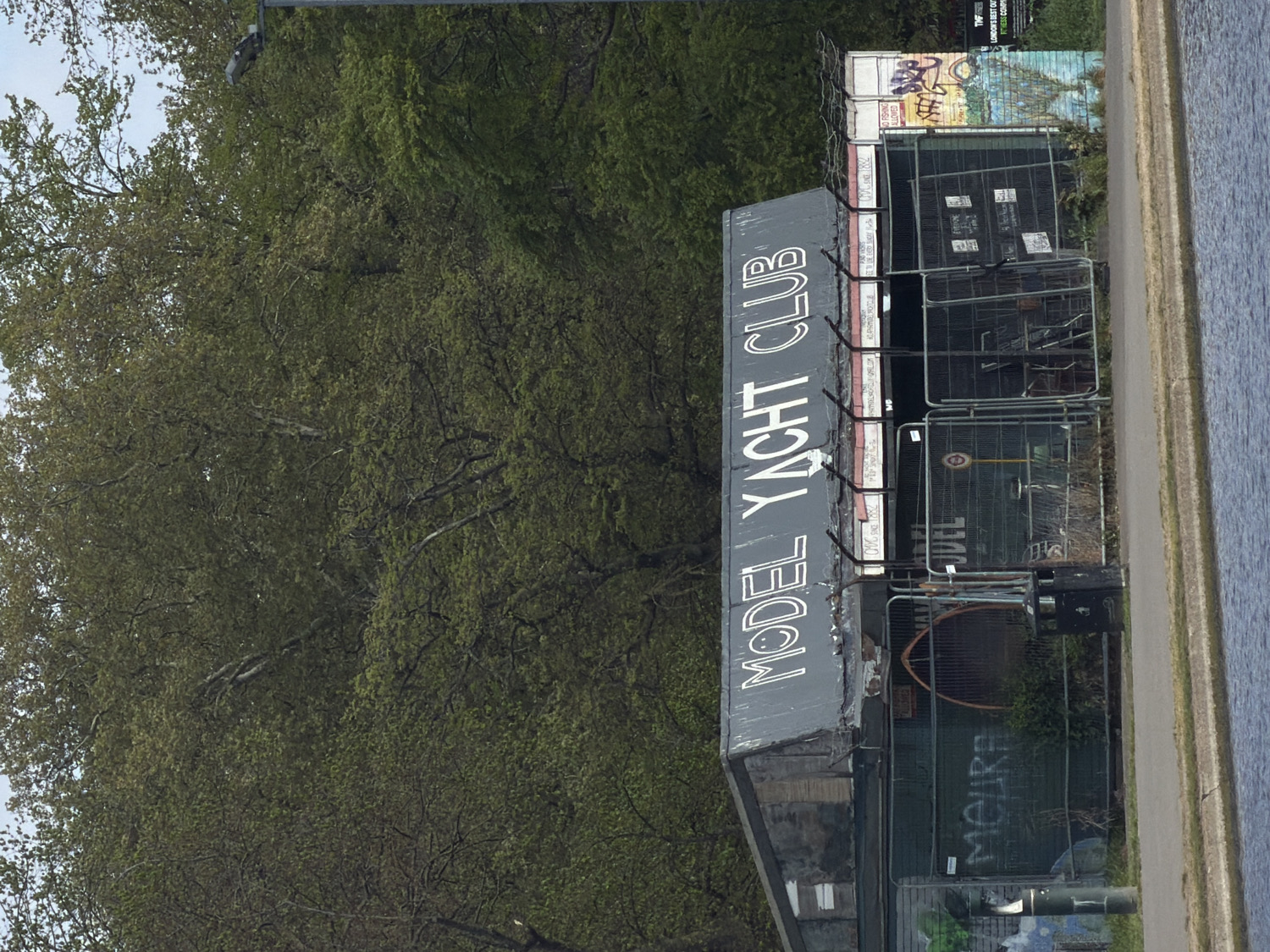The image captures an outdoor scene featuring a building, lush green trees, and a waterway under an overcast sky. The photograph is oriented vertically, showing the building horizontally.
The central subject is a dark-colored, rectangular building with large, white capital letters spelling "MODEL YACHT CLUB" prominently displayed on its side. The building appears somewhat weathered, with some peeling paint and signs of age.
To the left of the building (as viewed in correct orientation), a dense canopy of green trees fills a significant portion of the frame, suggesting a park or forested area adjacent to the scene. To the right, a paved path runs alongside a calm body of water, likely a canal or river, indicating a towpath or riverside walk. The water's surface reflects the light and appears serene.
There are metal fences, possibly temporary construction barriers, positioned in front of and around parts of the "MODEL YACHT CLUB" building. These fences have several signs attached, some of which are illegible, while others show various graffiti tags. One visible piece of graffiti on a surface behind the fences reads "MOURA." In the background, behind the fences, another structure or wall is visible with more graffiti, featuring vibrant colors and distinct lettering. A lamppost or street light pole is visible at the top edge of the image, extending into the tree line.
No direct human activity is observed, leaving the scene tranquil and static. The natural light suggests it is daytime, possibly late morning or afternoon, with soft, diffused light characteristic of an overcast day. Given the provided context, the scene is located in London, United Kingdom, likely along one of its many canals or waterways. A poster near the top right, behind the fences, contains text like "TYF" and "LONDON."