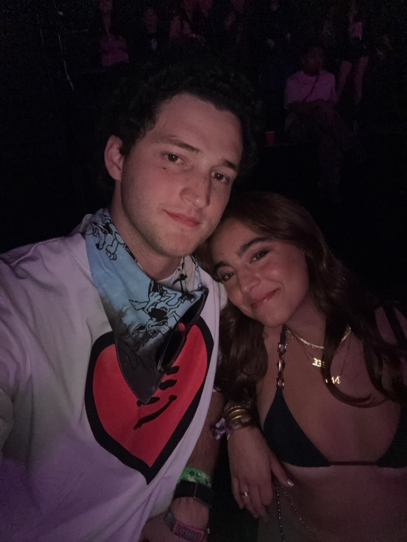 A medium shot captures a man and a woman posing closely, possibly in the city of Indio, United States. The man, positioned on the left, has dark, curly hair and is wearing a white T-shirt adorned with a large red heart and a light blue bandana with cartoon-like figures. He is also wearing several wristbands. Sunglasses are attached to the bandana. To his right, the woman has long, brown hair and is wearing a dark bikini top and layered necklaces. She is also wearing bracelets on her wrist and a ring on her finger.
They are in what seems to be a crowded area with other people faintly visible in the background. The overall lighting is dim, indicating the scene is likely indoors or at night.