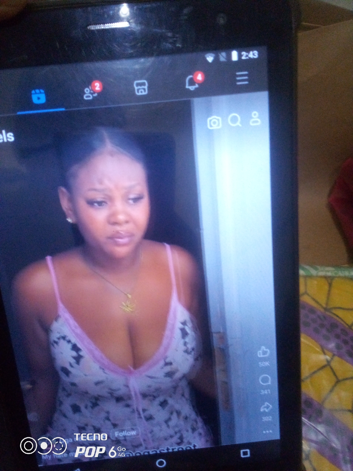 The image shows a close-up of a smartphone screen displaying a video. On the screen, a young woman with dark hair is visible, wearing a light-colored, patterned dress with pink trim. She is wearing a necklace. The phone's interface elements are also visible, including icons for Reels, people with a notification count of "2", a notification bell with a count of "4", a menu icon, a camera icon, a search icon, a user profile icon, a "like" icon with "50K", a comment icon with "341", a share icon with "302", and the text "TECNO", "Follow", "POP 6 Go 4G". The background outside the phone screen is out of focus and appears to be an indoor setting with a patterned surface and some fabric. The time on the phone's status bar reads "2:43". There is no identifiable location within the city of Howayi, Nigeria.
