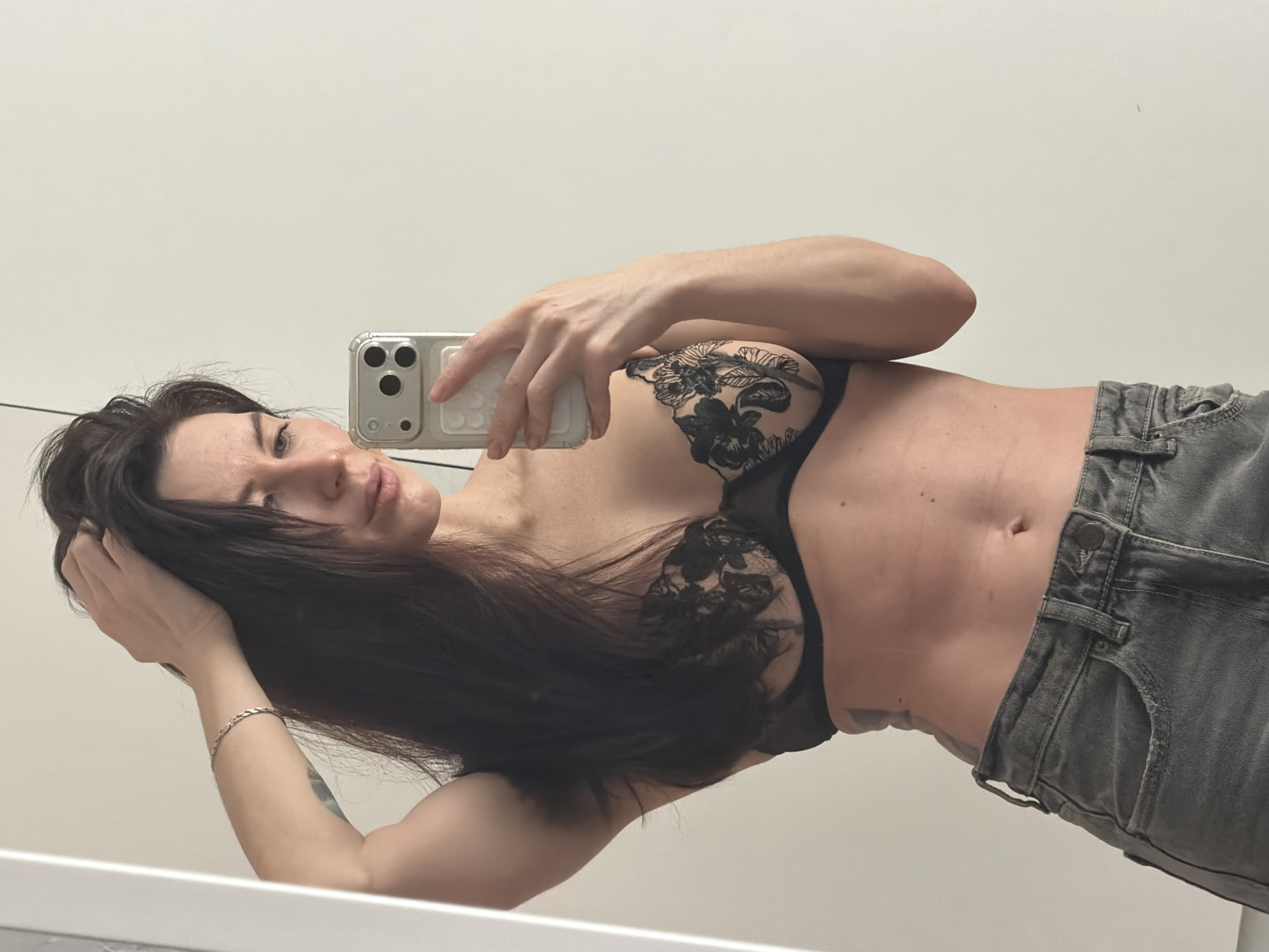 A woman with long dark hair, wearing a black lace bra and gray jeans, is taking a selfie. She is holding a silver iPhone with her right hand, and her left hand is behind her head. She is standing in front of a white wall. There is a silver bracelet on her left wrist. There is no evidence to suggest that this image was taken in the city of Perm, Russia.