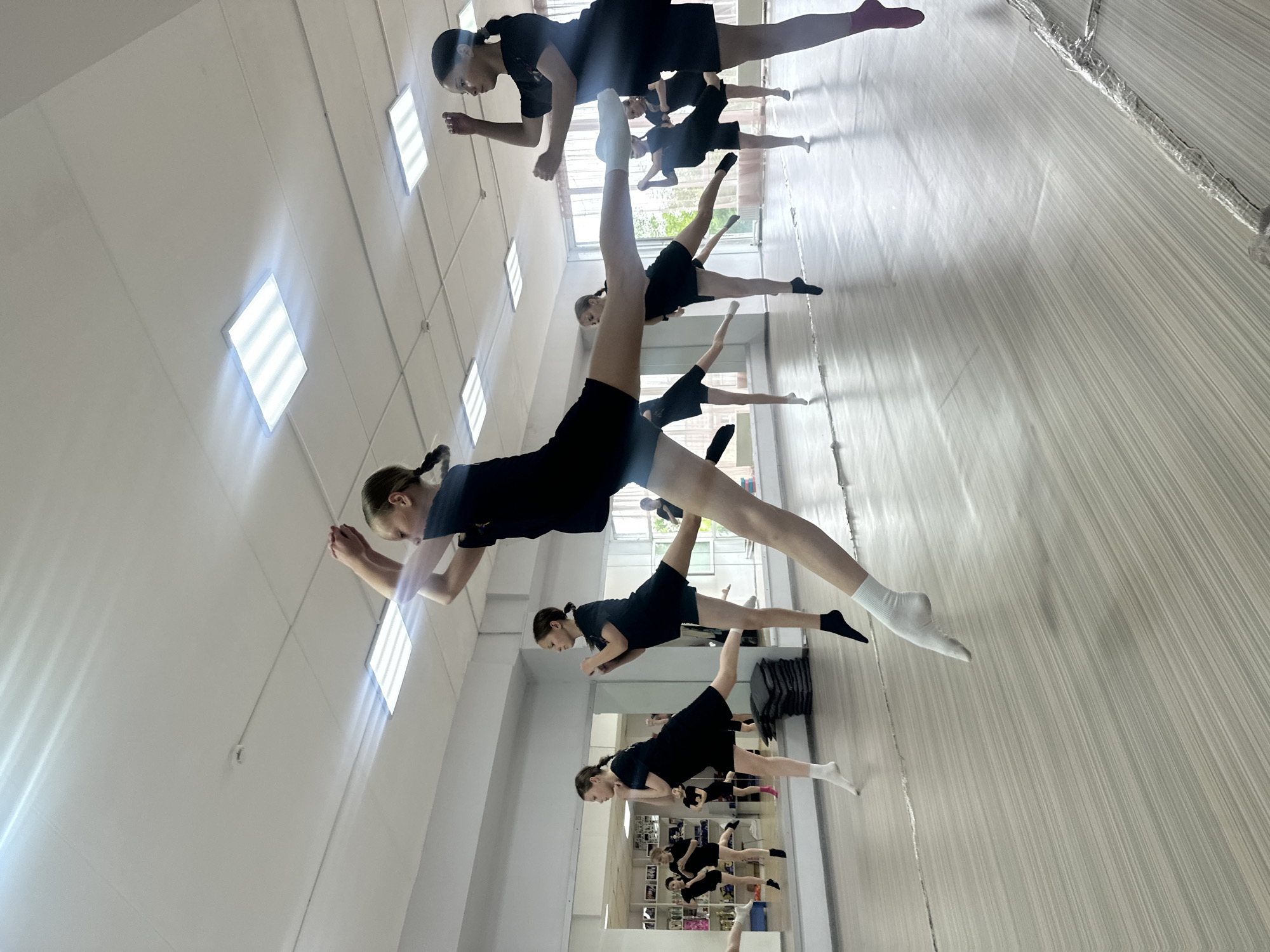 In a dance studio located in Votkinsk, Russia, several young girls in black dance attire are captured mid-air performing balletic jumps. The studio features a polished wooden floor, a large mirrored wall reflecting the dancers, and fluorescent lighting fixtures on the ceiling. The girls are positioned at various points in their jumps, with one central figure in the foreground executing a high leap with one leg extended. The overall scene depicts a moment of synchronized athletic movement and focused practice.