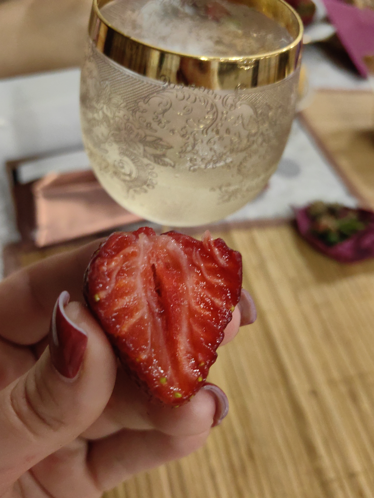 A close-up shot shows a person's hand holding a halved strawberry. The strawberry is bright red with visible seeds and a textured interior. The person's fingernails are painted a deep red. In the background, a glass with a gold rim is filled with a light-colored beverage, possibly wine or champagne. The glass has an intricate etched design. Also in the background, a portion of a pink paper object and a small dish with some food are visible. The scene appears to be set indoors, possibly in a restaurant or cafe, with a wooden surface in the foreground. There is no specific information within the image to determine the location in the city of Sochi, Russia.