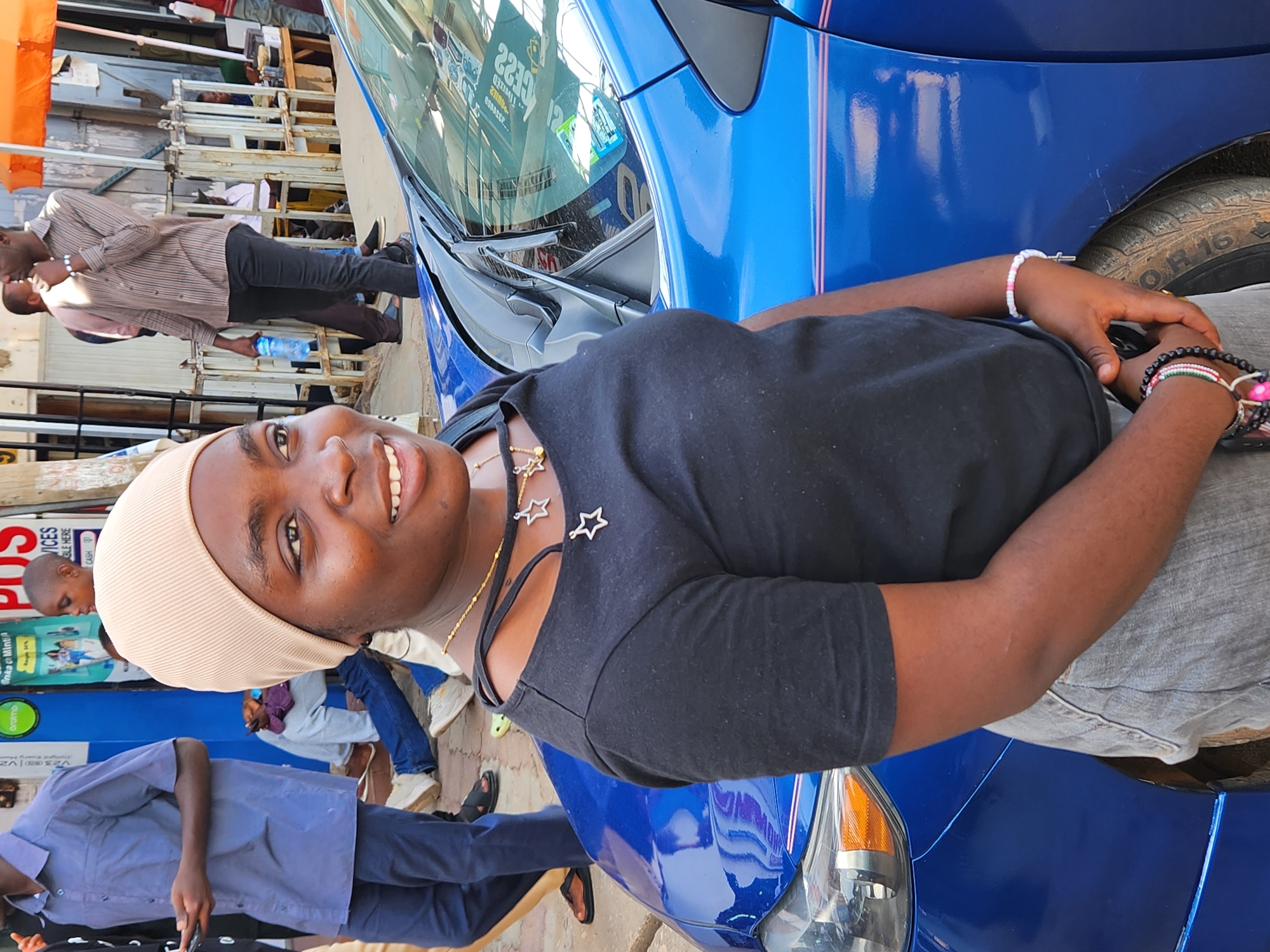 This full-color, daylight photograph captures a bustling urban street scene, identified as Jos, Nigeria. The image is oriented with the main subject's head towards the top-left, suggesting it was originally taken in portrait mode and then rotated.
In the foreground, a person with dark skin is prominently featured, smiling broadly at the camera. They are wearing a light-colored beanie or head covering, a black short-sleeved top with cut-out shoulder details, and a gold-colored necklace with star-shaped pendants. Multiple bracelets, featuring beads in white, pink, black, green, and red, adorn both wrists, and their hands are clasped in front of them. This individual appears to be leaning against or positioned beside a vibrant blue car, which dominates the midground and lower portion of the image. The car has subtle pink pinstripes along its side, and reflections on its windshield show glimpses of the background. A partial view of the car's tire reveals "OR 16".
The background is lively, featuring several other people and commercial elements. One man in a striped long-sleeved shirt and dark pants is visible, holding a blue water bottle. Another individual in a blue short-sleeved shirt and dark pants is partially obscured. Further back, shop fronts or market stalls with metal frameworks are visible, suggesting a commercial area.
Visible text on various signs includes "SERVICES", "POS", "CIRCLE HERE", and "CASH". Another green and blue sign partially shows "oromo". The bright lighting indicates a sunny day. The main subject's cheerful expression conveys a relaxed and happy mood amidst the everyday activity of the street.