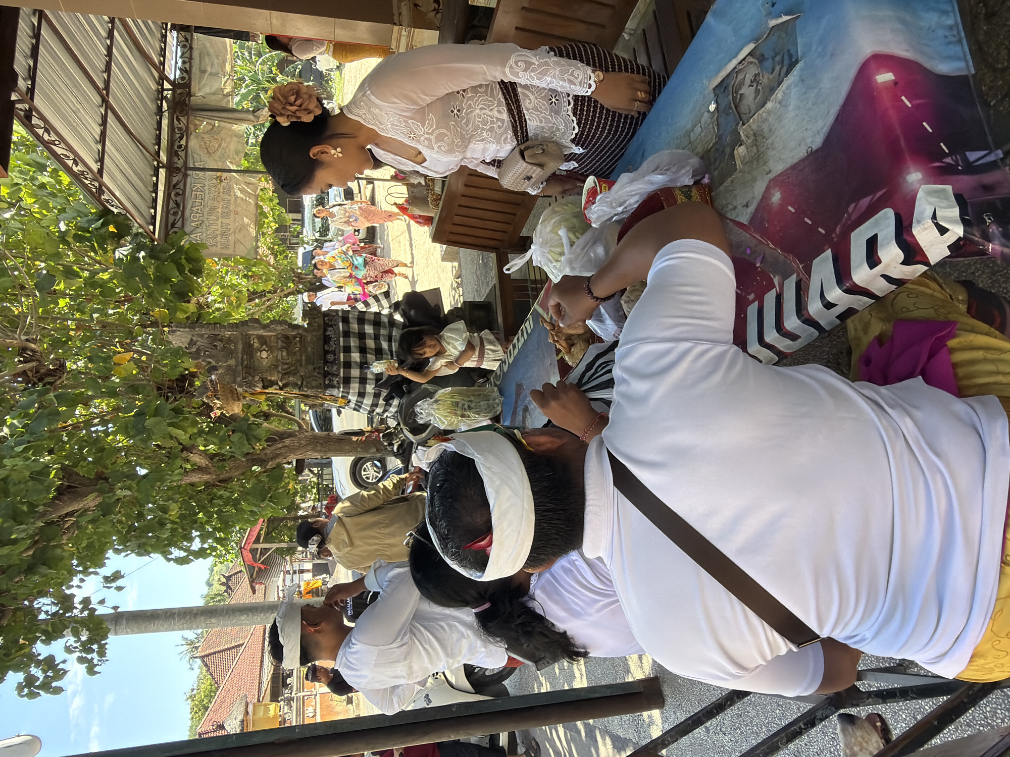 This image depicts an outdoor scene in Nusa Penida, Indonesia, likely near a temple or communal gathering area, given the traditional attire and the presence of offerings. In the foreground, two individuals, a man and a woman, are seated at a table covered with a colorful, torn tablecloth. The man, viewed from the back, wears a white shirt and a white head covering. The woman, in the middle ground, is also wearing a white, intricately embroidered top and a dark, striped sarong, with a flower in her hair. She is interacting with a small child who is reaching towards the table.
Several other people are visible in the background, some wearing traditional Balinese white clothing and headbands, suggesting a cultural event or religious ceremony. They are standing near buildings with traditional tiled roofs and a paved road. Trees with dense green foliage are prominent, providing shade. The overall atmosphere appears to be one of community gathering and observance.