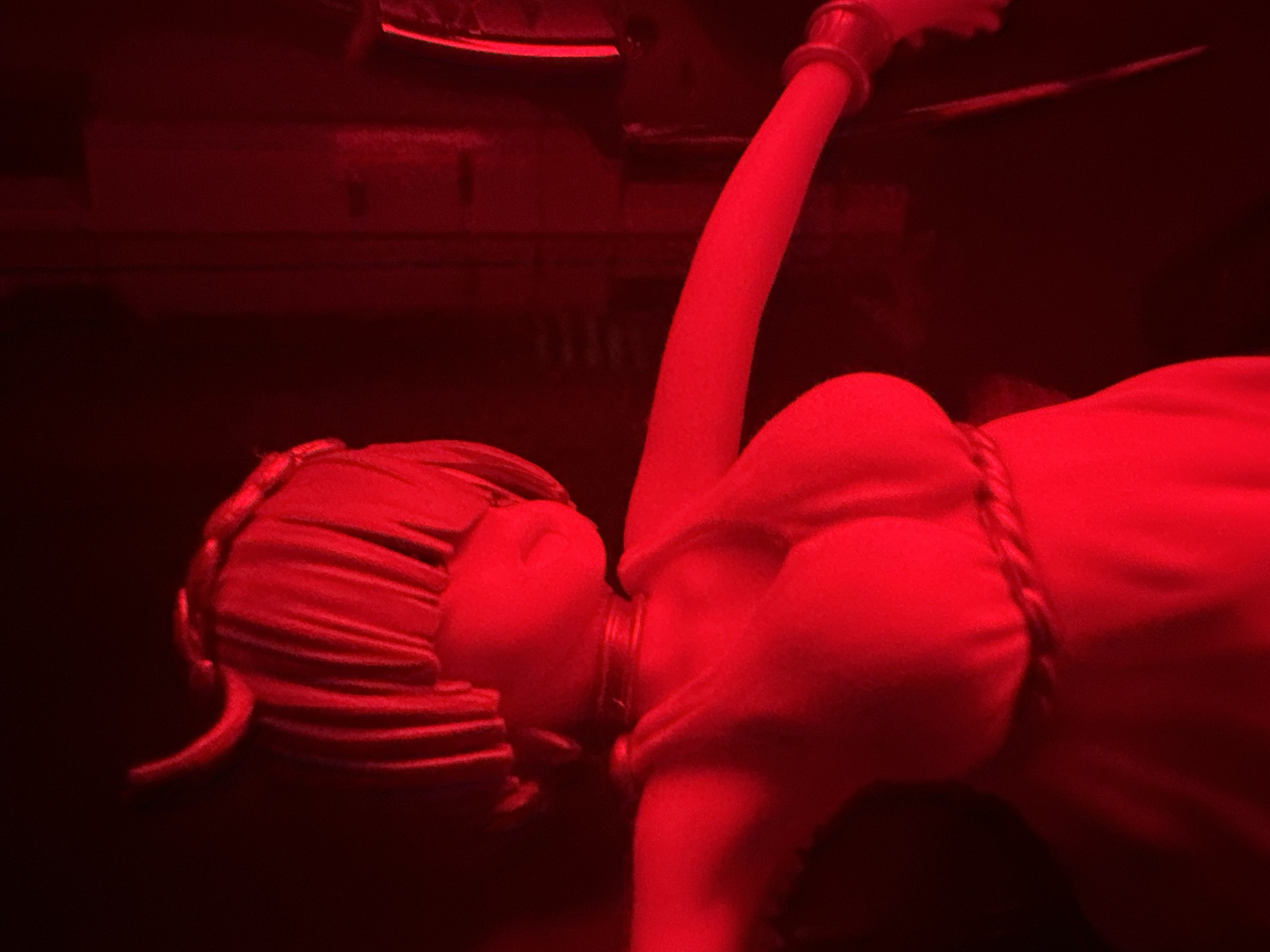 A full-body shot captures a doll with red lighting. The doll appears to have devil horns and a bob haircut. It wears a red dress and a necklace. The background is dark, with some blurry shapes that could be furniture or walls.