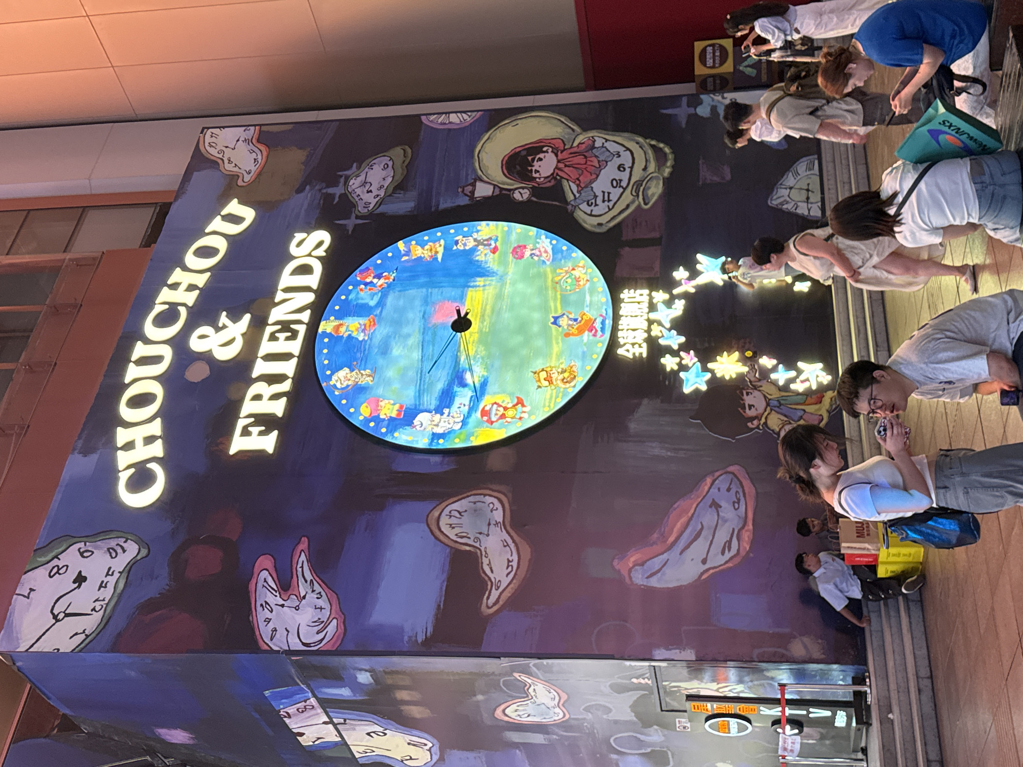 The image captures a vibrant nighttime scene in Shanghai, China, likely on a busy street or plaza. The focal point is a large, illuminated mural adorning the side of a building, featuring a surrealist theme with melting clocks, reminiscent of Salvador Dalí's work. The mural displays the text "CHOUCHOU & FRIENDS" in glowing golden letters. A large, round clock face is integrated into the mural, adorned with various cartoon characters. Below the mural, a diverse group of people are visible. Some are standing and engaged in conversation, while others are seated on benches. Two young women in the foreground are looking at their phones. In the mid-ground, a few individuals are walking, and one person is sitting near some shopping bags, one of which is branded "MUJI." The environment appears to be an urban setting, with building facades visible in the background, some with windows and architectural details. The lighting is a mix of artificial illumination from the mural, streetlights, and possibly building lights, creating a lively and dynamic atmosphere.