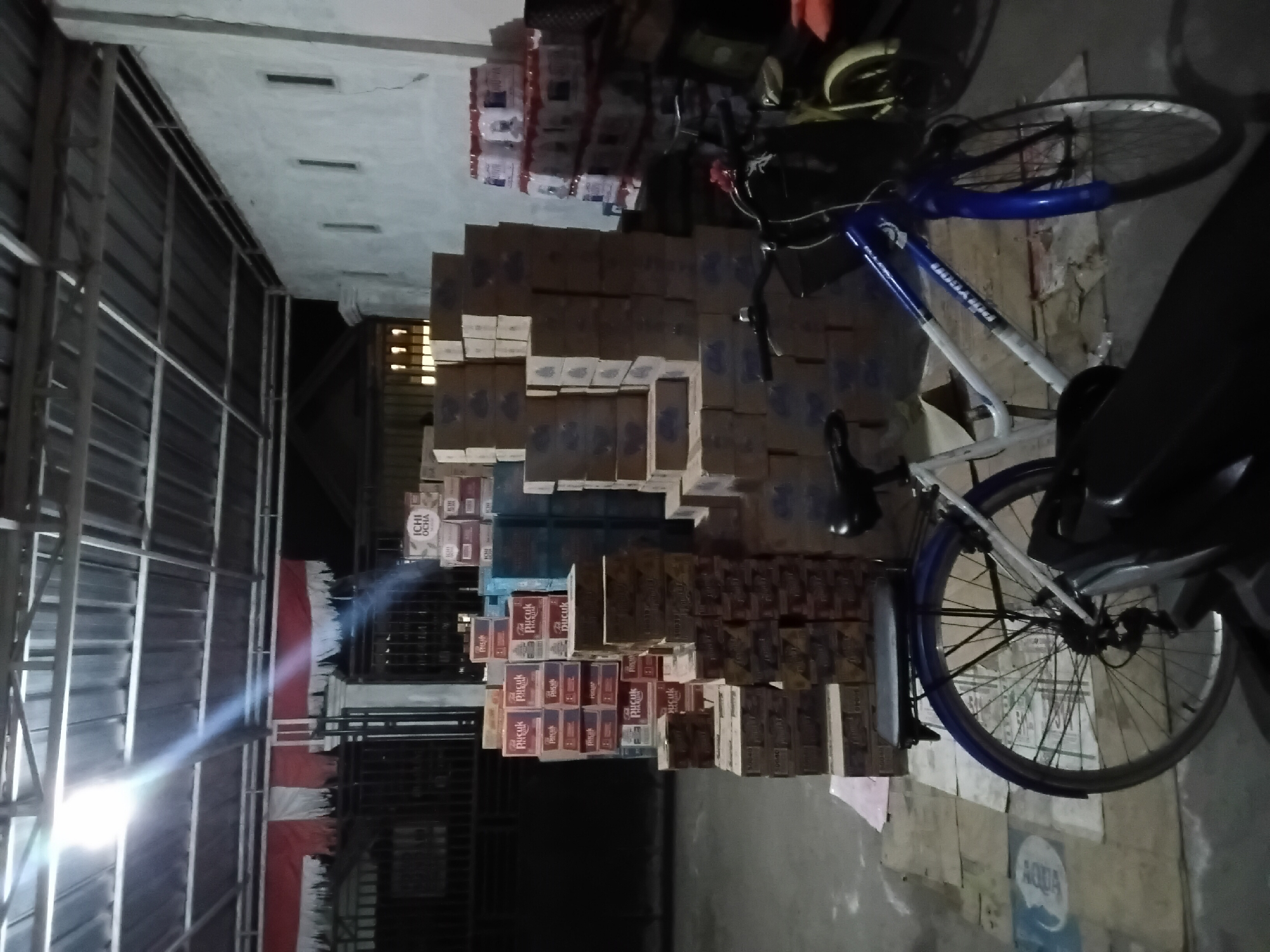 In the city of Trenggalek, Indonesia, a dimly lit warehouse or storage area is depicted. The space is filled with stacks of cardboard boxes, some appearing to be beverage containers. Prominently featured in the foreground are two bicycles. One is a white and blue bicycle with a black seat, and the other, partially obscured, appears to be a yellow or lime green bicycle with a black seat. The bicycles are positioned on a floor covered with what looks like flattened cardboard boxes and some scattered paper or fabric. The environment has a corrugated metal roof supported by metal beams. A national flag of Indonesia, red and white, hangs near a gated entrance to the left. The lighting is artificial, with a strong, direct beam of light originating from the upper left, casting shadows and creating lens flare effects.