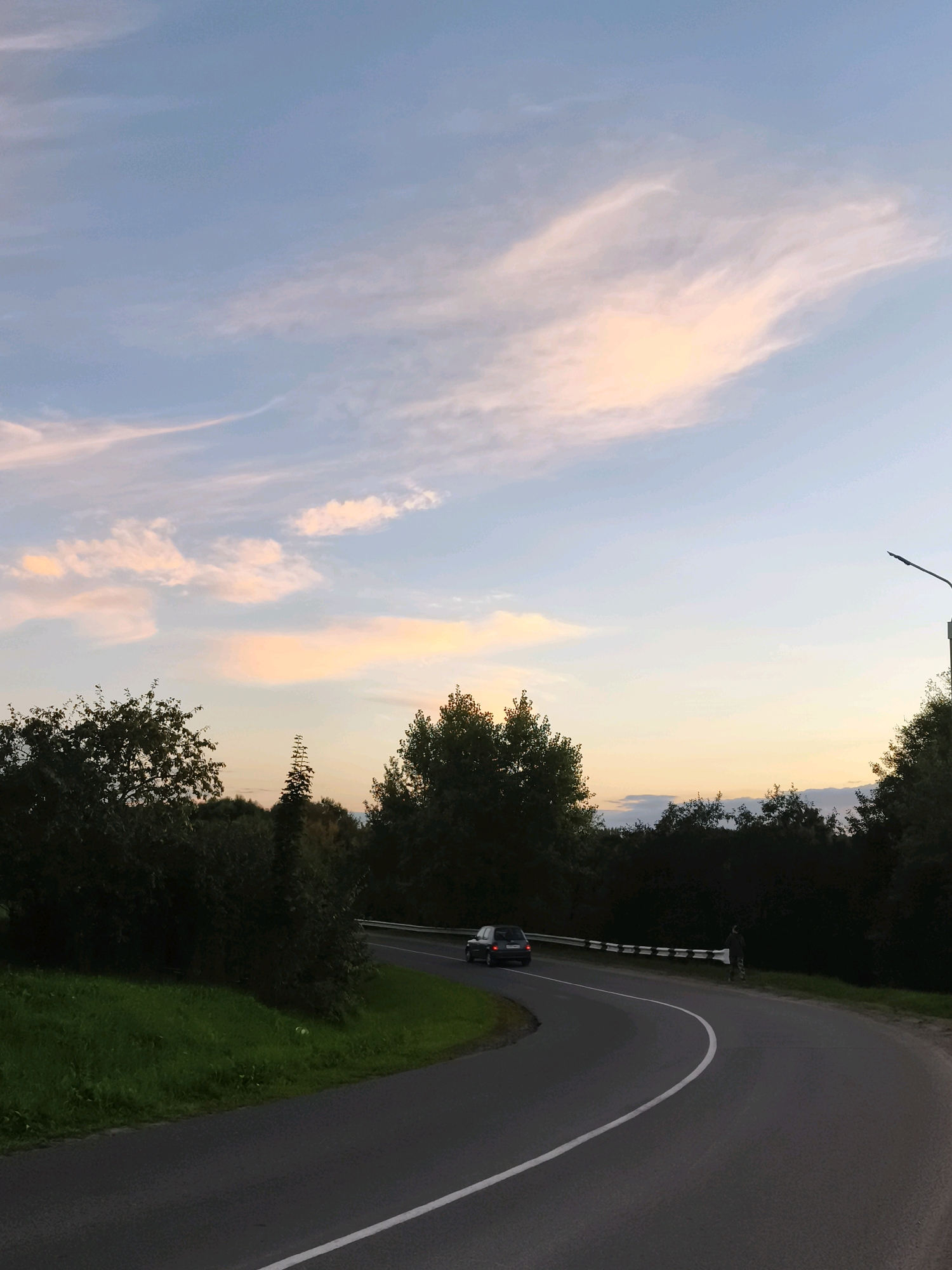 The image captures a winding asphalt road through a wooded area, likely in the vicinity of Горскі сельскі Савет, Belarus, during twilight. A dark-colored car is driving on the road, navigating a curve. The sky above is a soft gradient of blue and pale pink, with wispy clouds illuminated by the setting sun. Dense green foliage lines the sides of the road, with trees forming a dark silhouette against the colorful sky. A streetlamp stands on the right side of the road. A figure, possibly a person, is visible standing near a guardrail on the right side of the road, facing away from the camera. The overall scene depicts a tranquil, rural landscape at dusk.